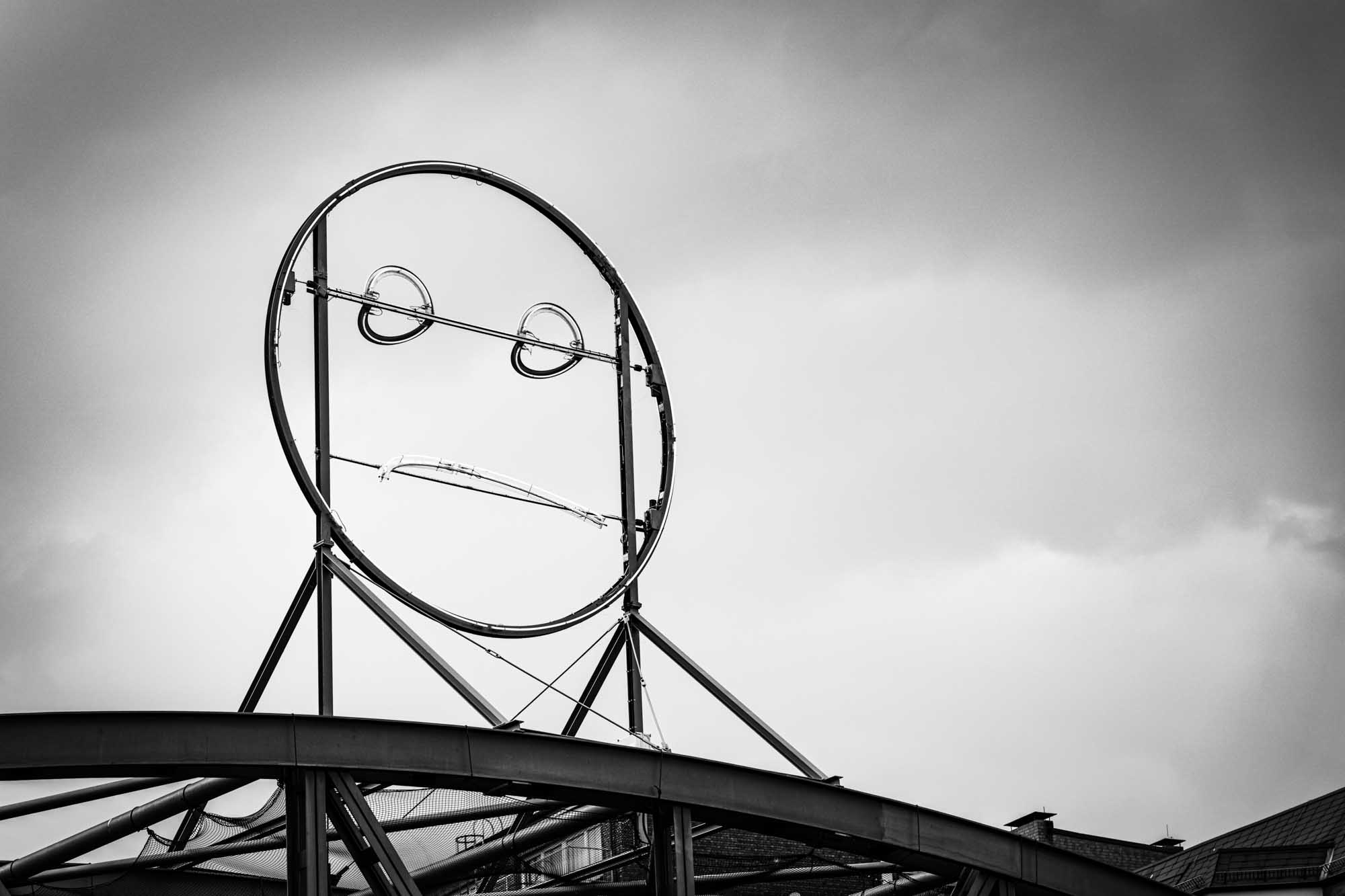 Modern metal sculpture of a face against a cloudy sky, featuring circular eyes and a line mouth.