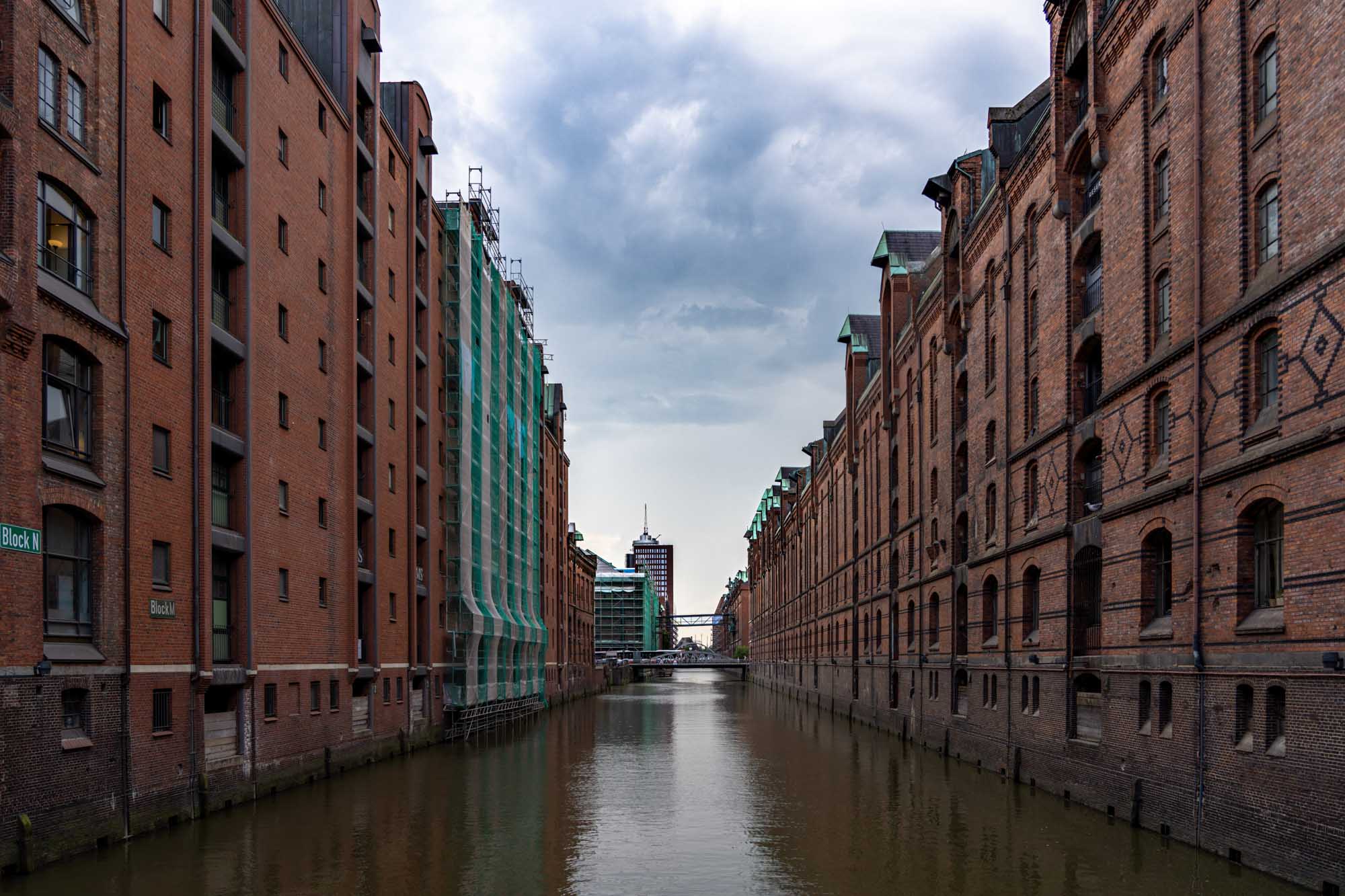 Historic brick warehouses flank a narrow water canal under a cloudy sky.