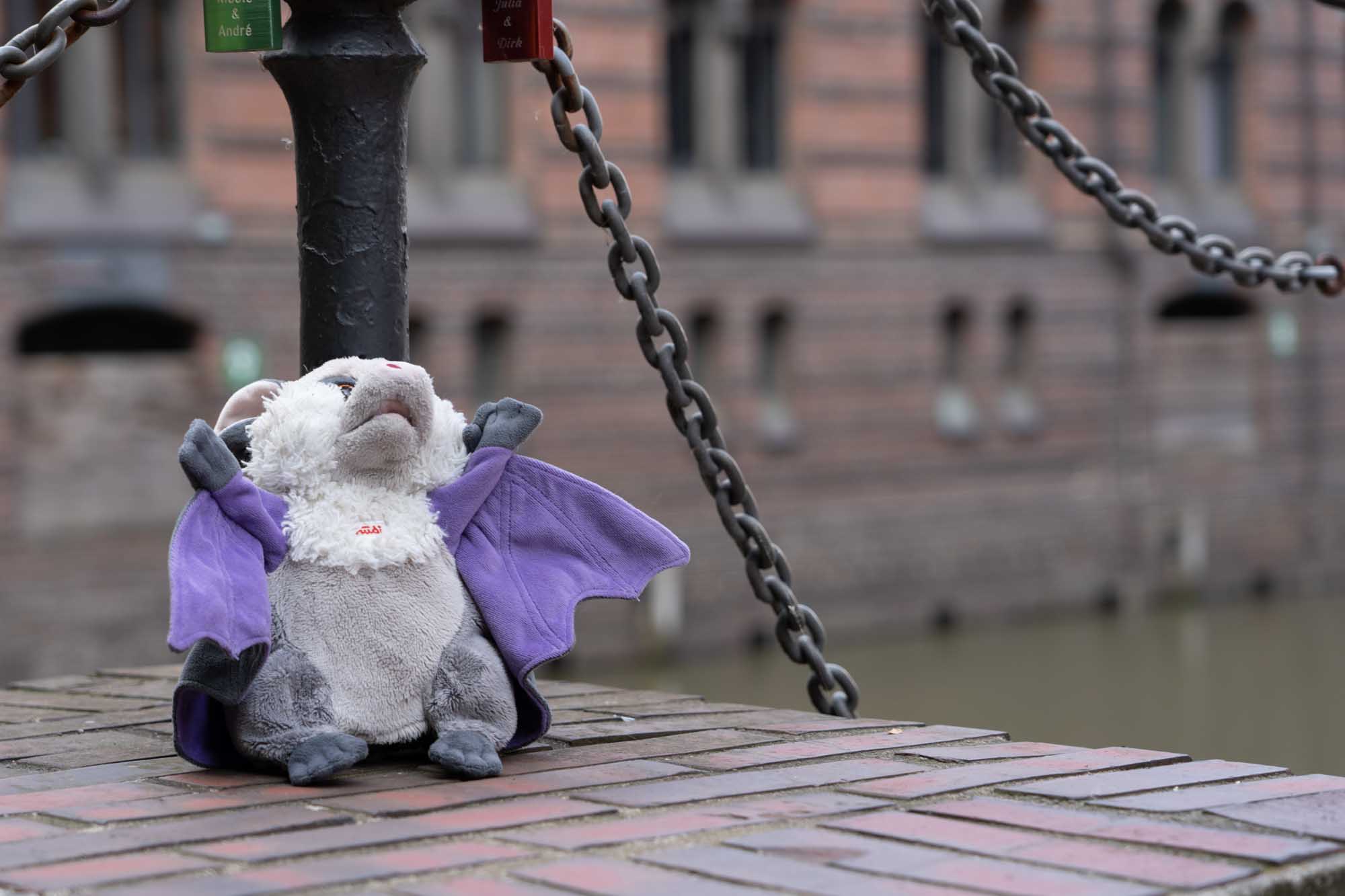 Stuffed bat toy with purple wings sits on a brick ledge near a pole with chains and love locks in an urban setting.