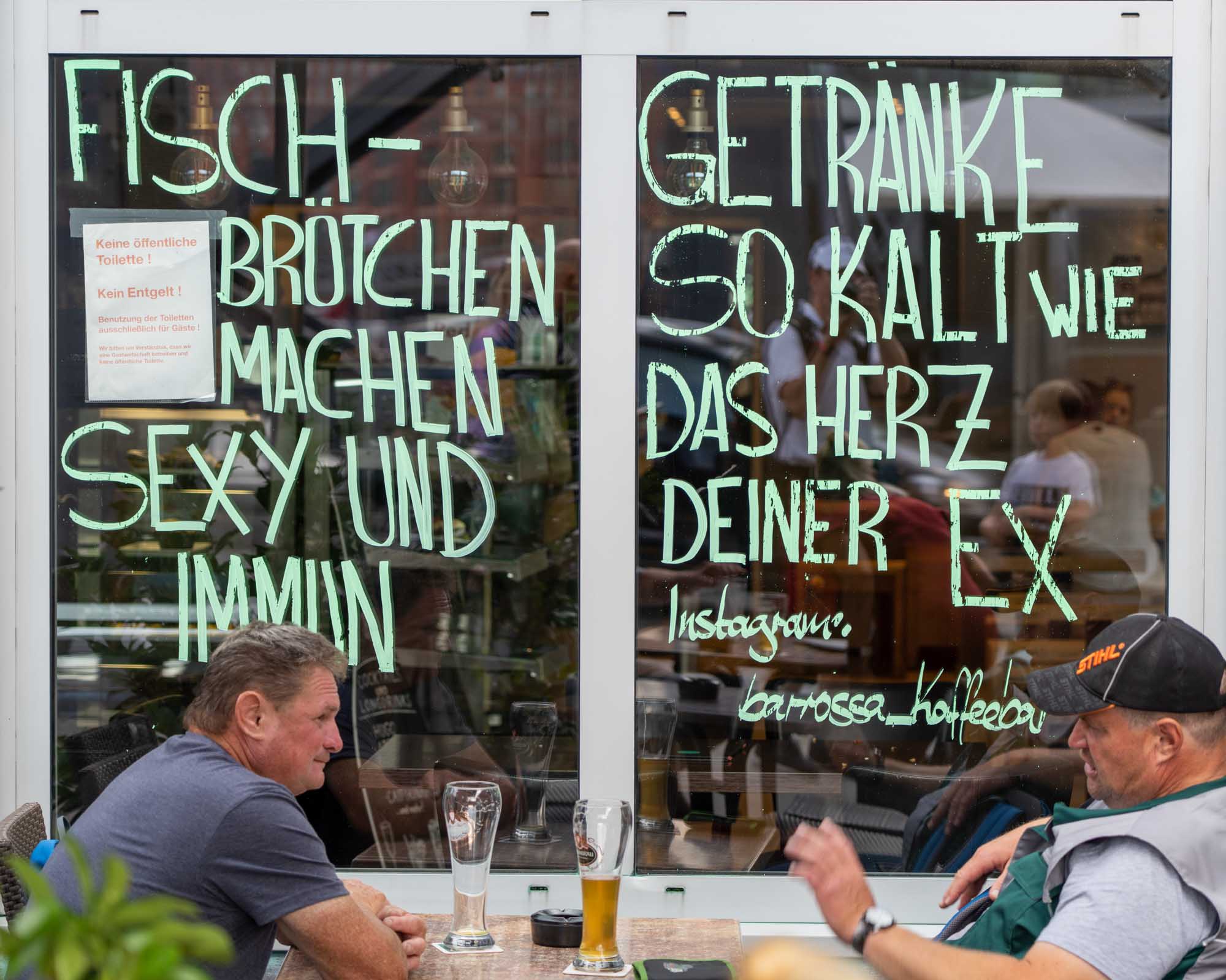 Two men sitting outside a café with humorous signs in German on the window behind them.
