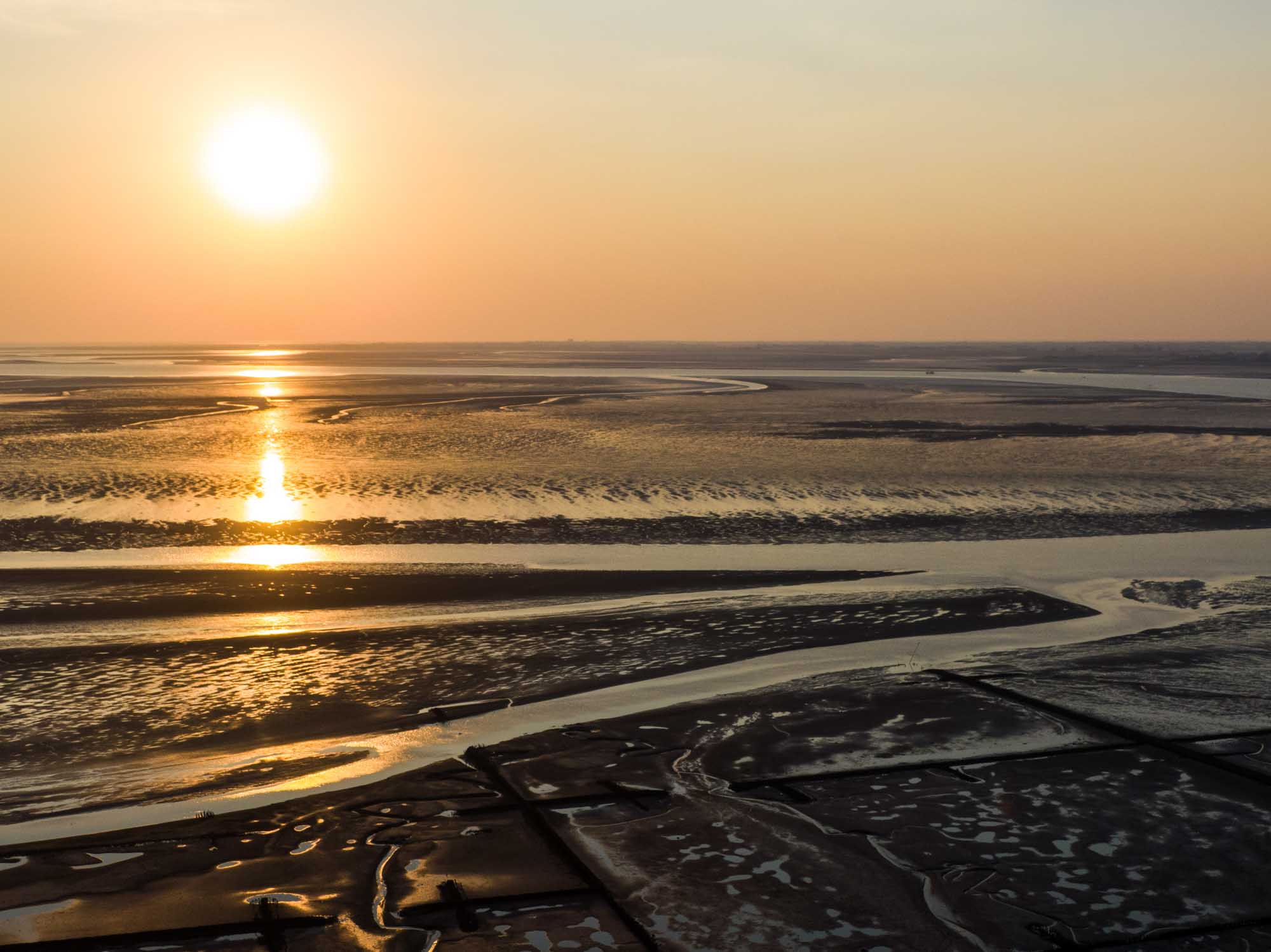 Sunset over expansive wetlands with reflective water channels and hazy horizon.
