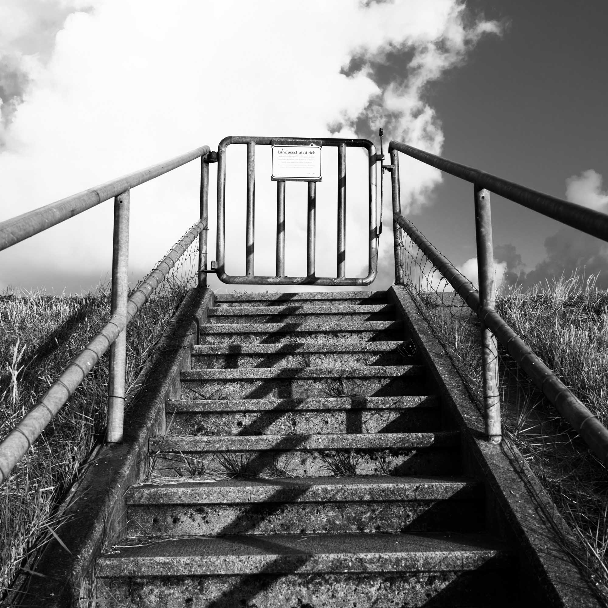 Concrete steps leading to a closed gate under a cloudy sky, flanked by metal railings in black and white.