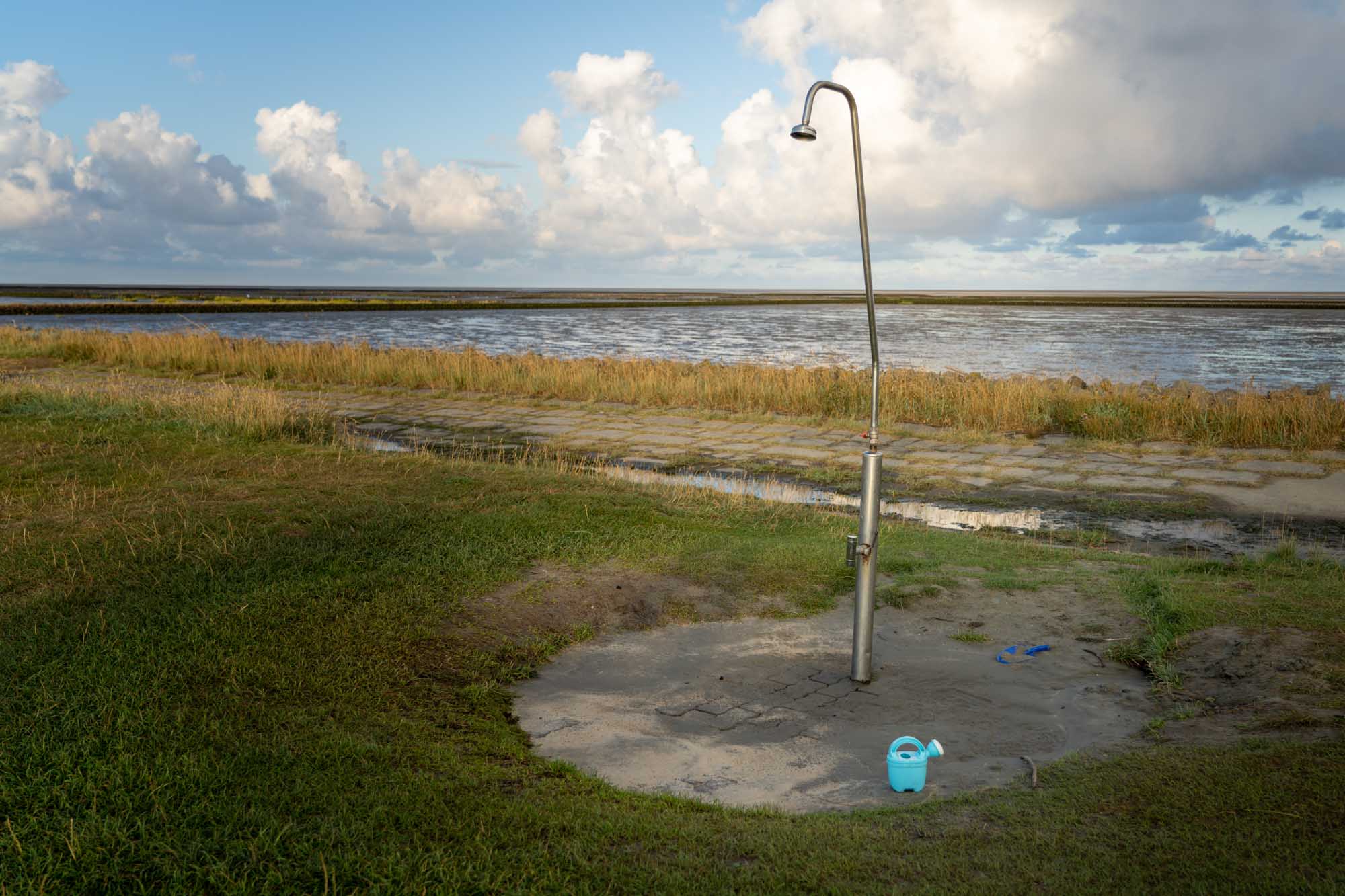 Outdoor shower near a coastal area with grassy surroundings and blue watering can.