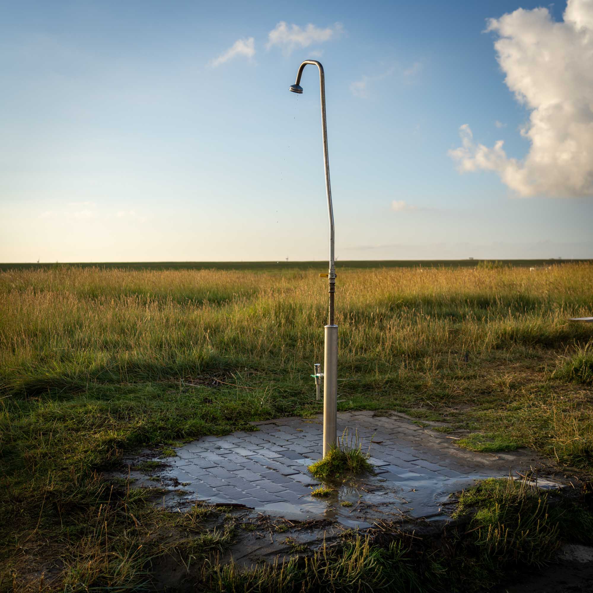 Outdoor shower in a grassy field, sunlight casting shadows on wet paving, with a blue sky and clouds overhead.