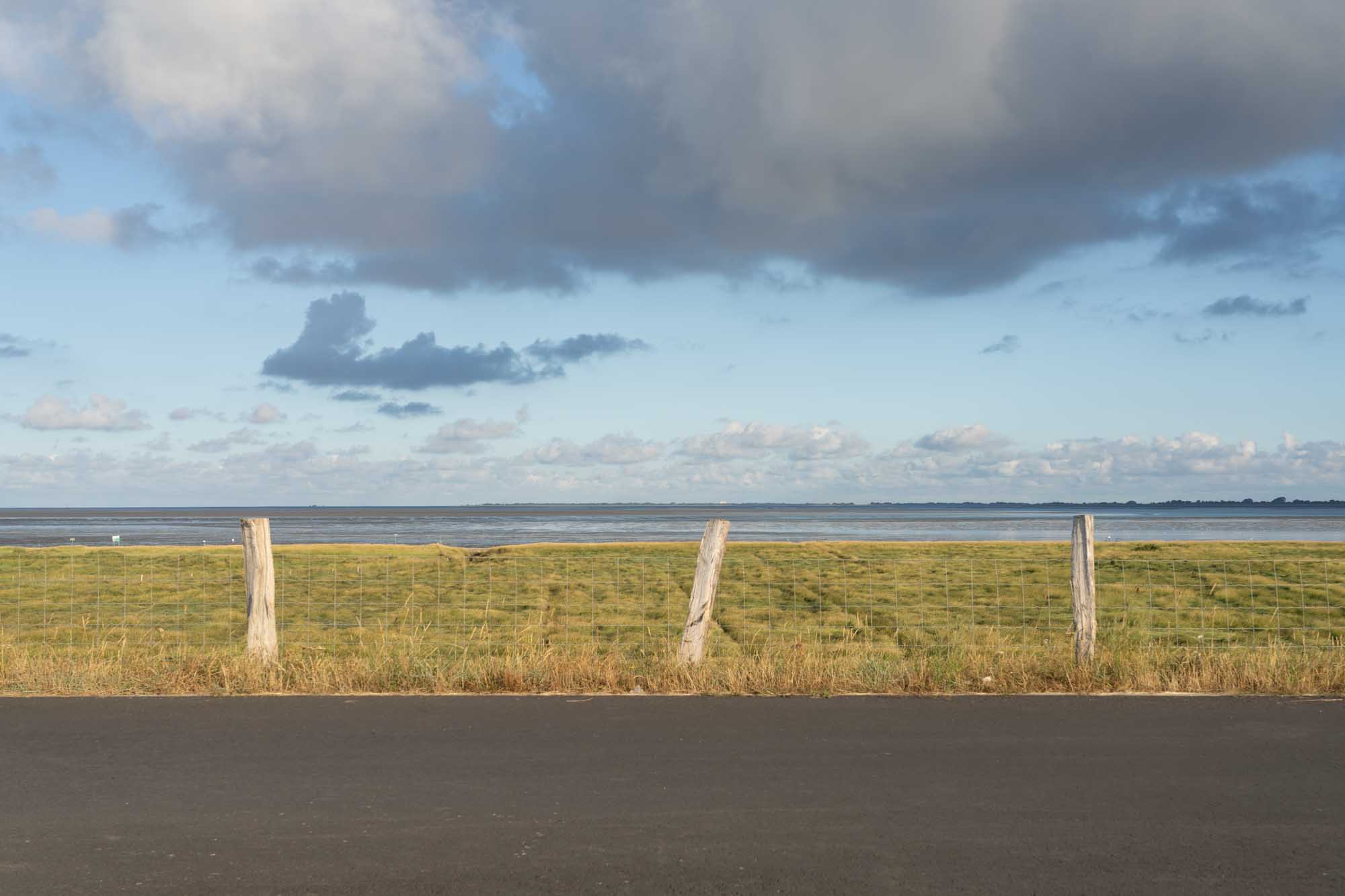 Coastal view with grassy field, wooden fence, and cloudy blue sky above a calm sea under sunlight.