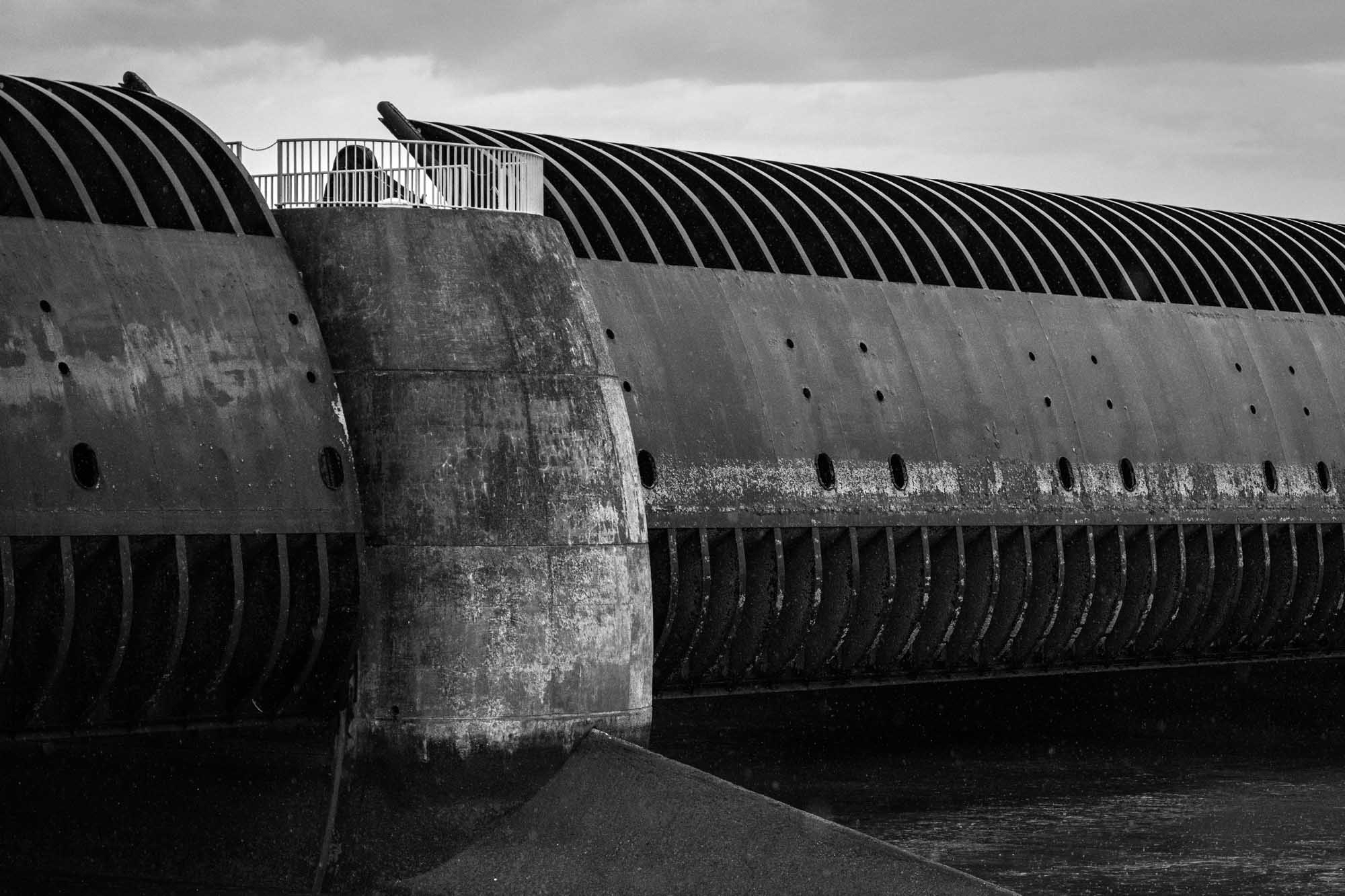 Black and white photo of large, curved industrial structure with dark vents and a viewing platform under cloudy skies.