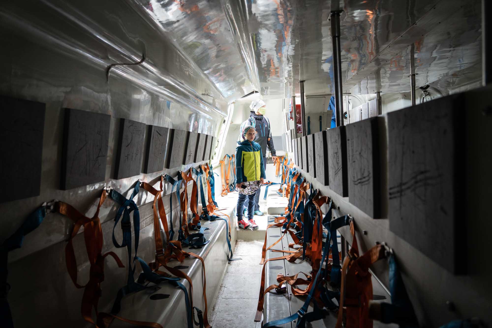 Child inside lifeboat with orange and blue harnesses, examining interior seating and equipment.