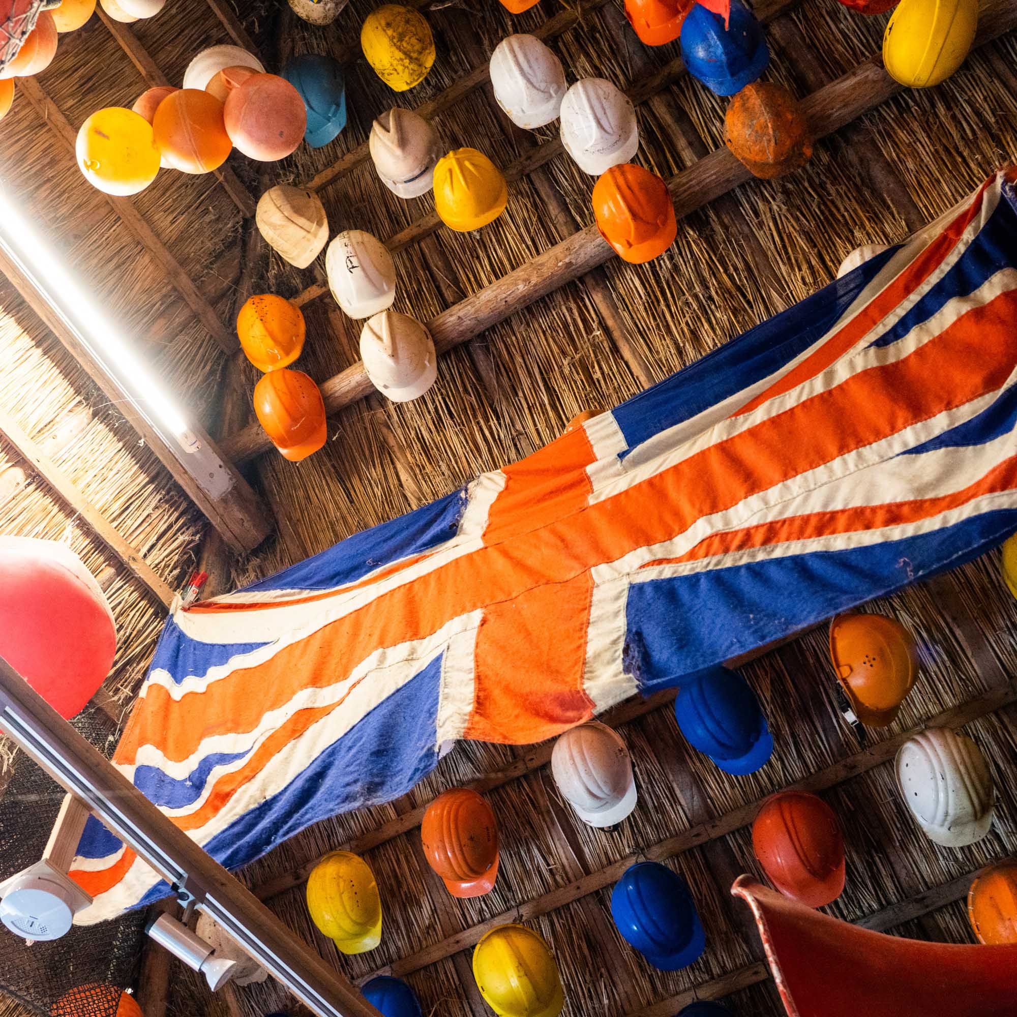 Colorful hard hats and Union Jack flag hanging from a rustic ceiling, creating a vibrant display.