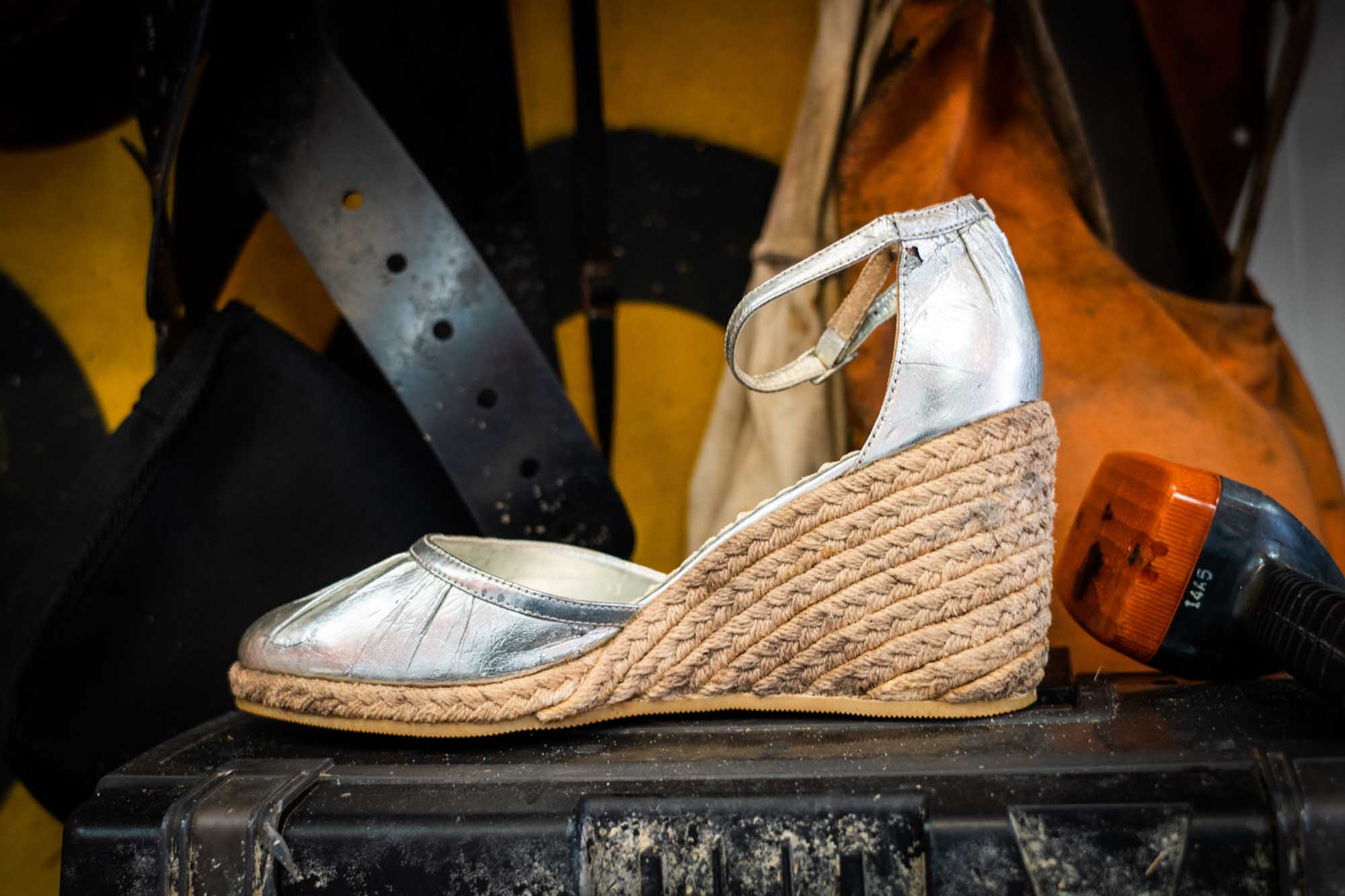 Silver espadrille wedge shoe with ankle strap, hand-sewn jute sole. Set against a rustic workshop background.