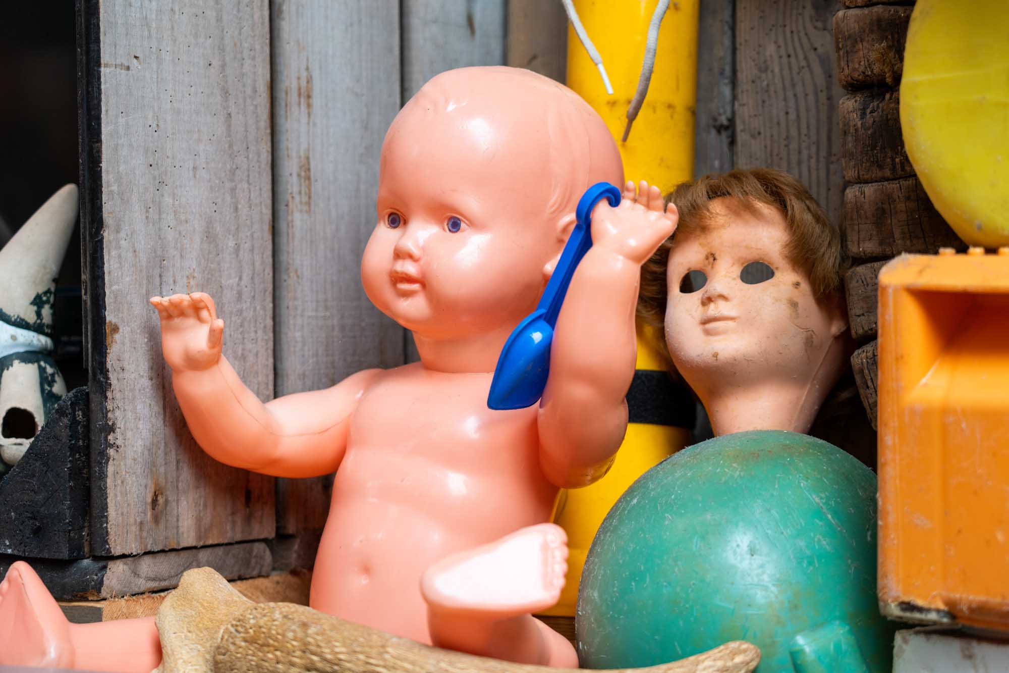 Plastic baby doll holding blue spoon next to weathered doll head and colorful toys in rustic setting.