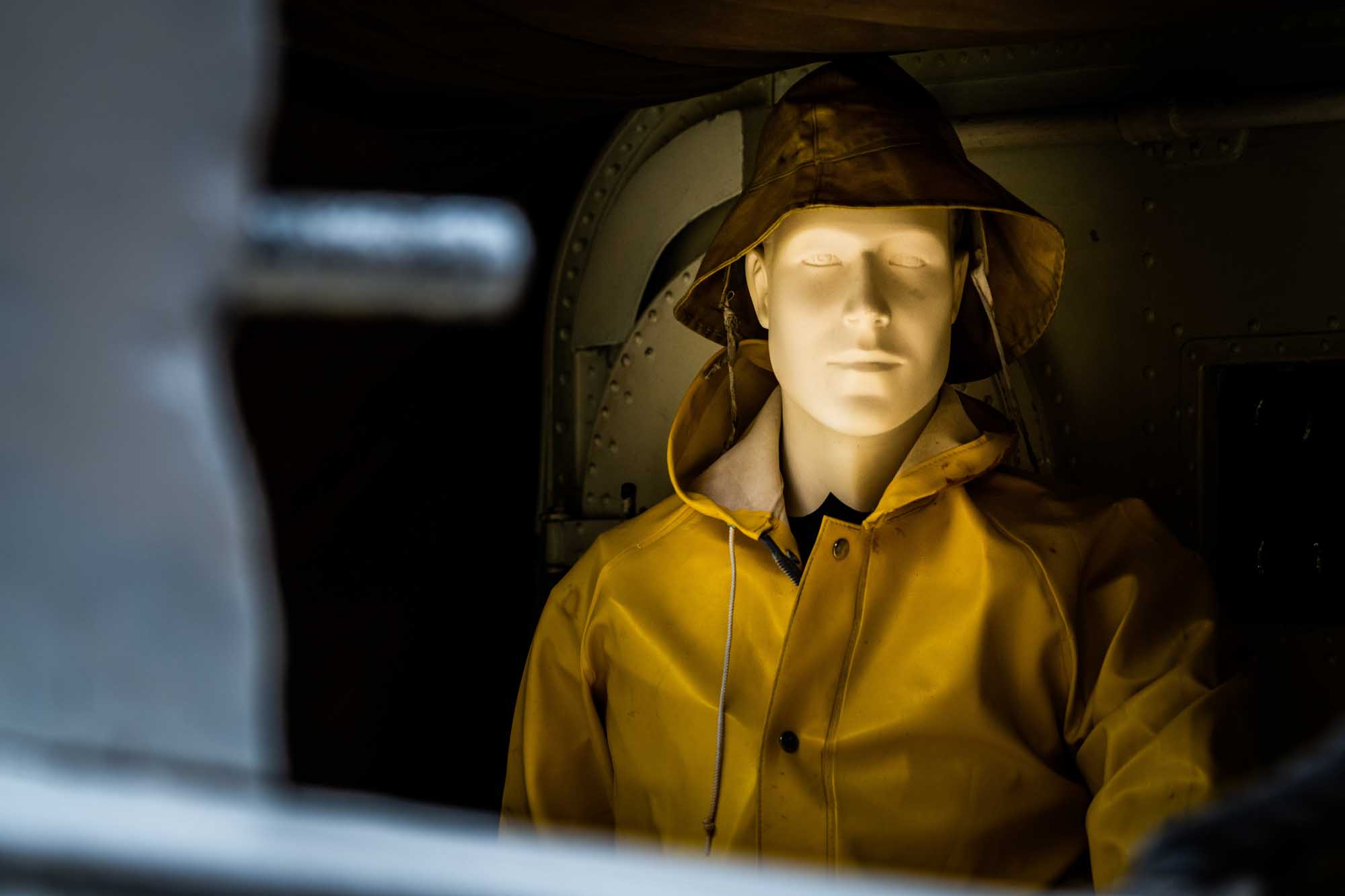 Mannequin in yellow raincoat and hat inside a dimly lit room, evoking a maritime or industrial theme.