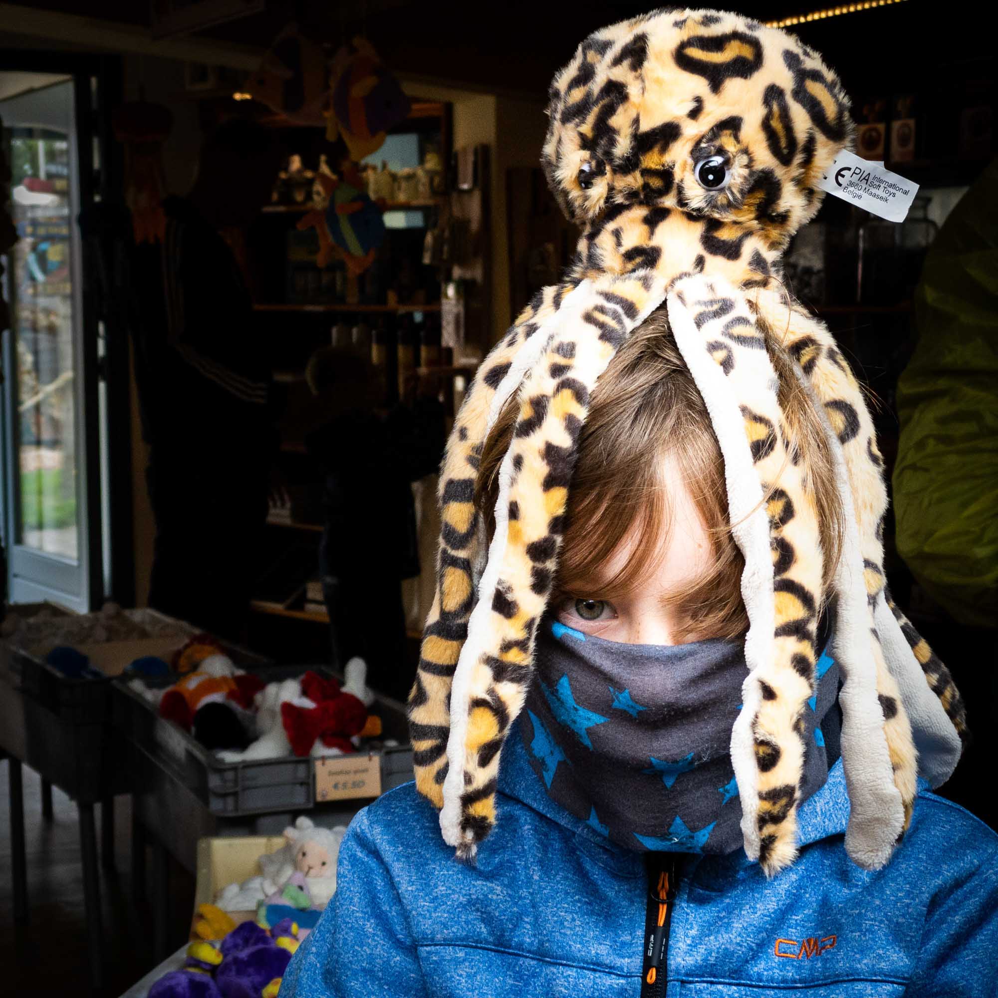 Child wearing plush leopard-patterned octopus hat and blue jacket in a toy shop.
