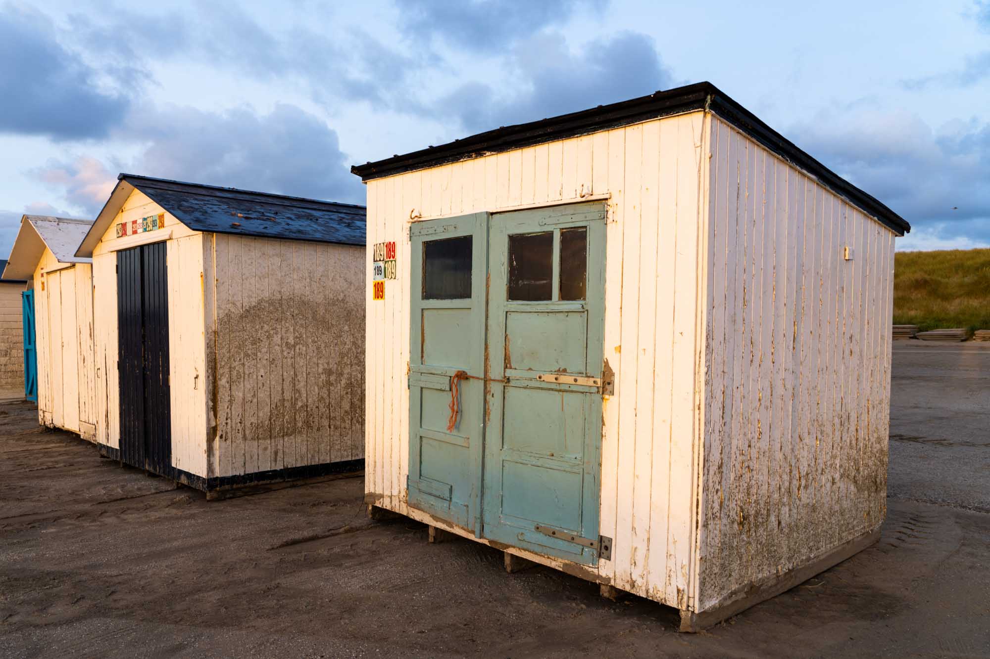Weathered beach huts with numbered signs and blue doors on a sandy shore under a cloudy sky.