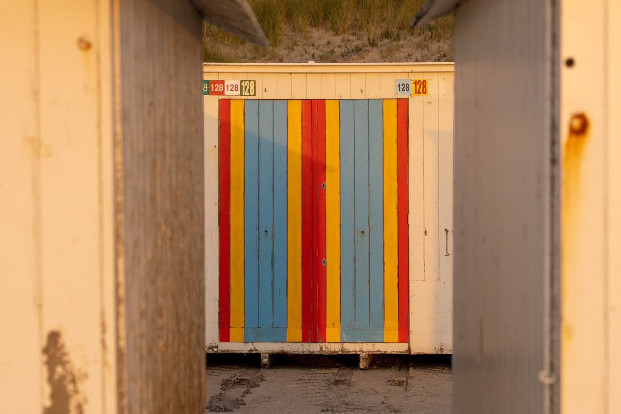 Colorful beach hut with red, yellow, and blue stripes framed by two wooden walls on a sandy shore.