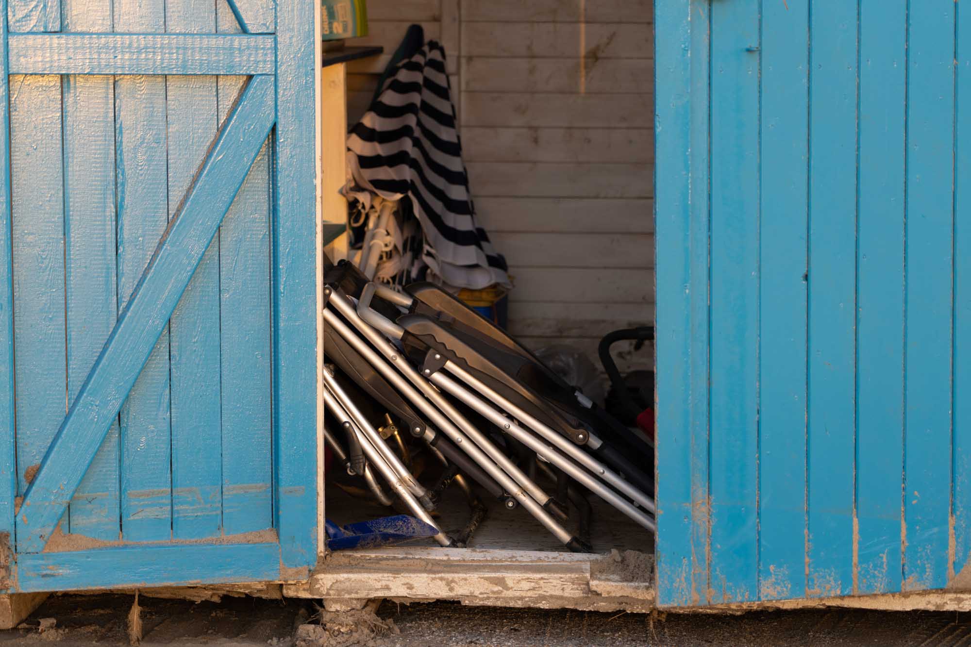 Blue wooden shed door slightly open, revealing folded chairs and striped fabric inside.