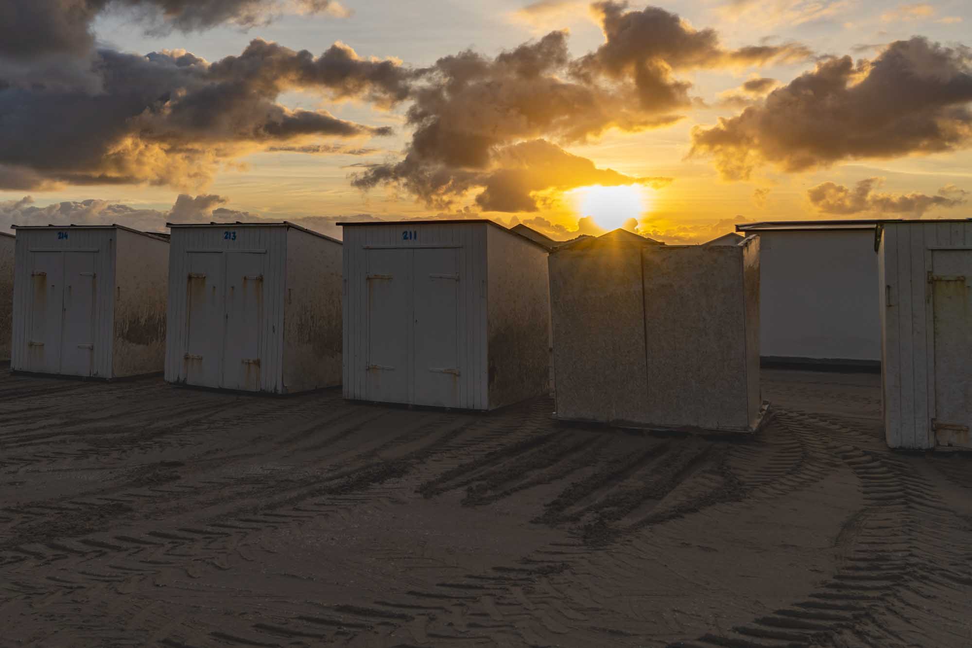 Beach cabins with numbered doors on sandy shore at sunset, dramatic clouds and sunlight creating a warm, serene atmosphere.