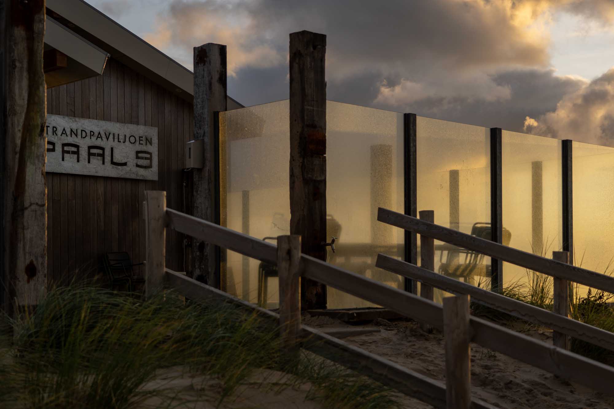 Beach pavilion entrance at sunset with wooden structures and glass panels, highlighted by dramatic clouds in the background.