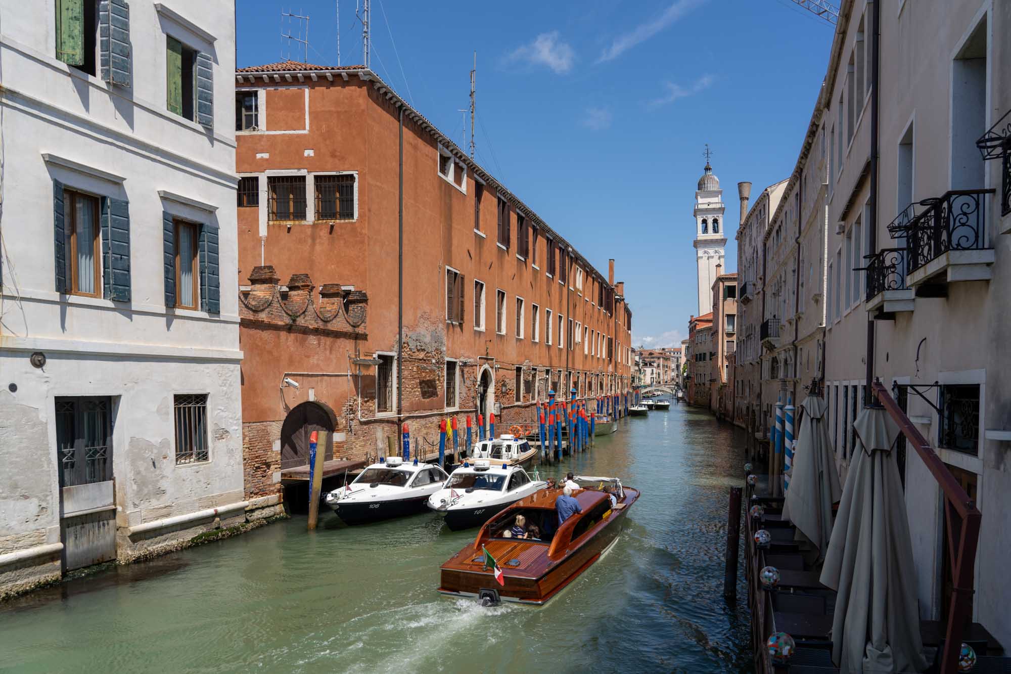 Water taxi on a Venetian canal with colorful buildings and a tower in the background under a clear blue sky.
