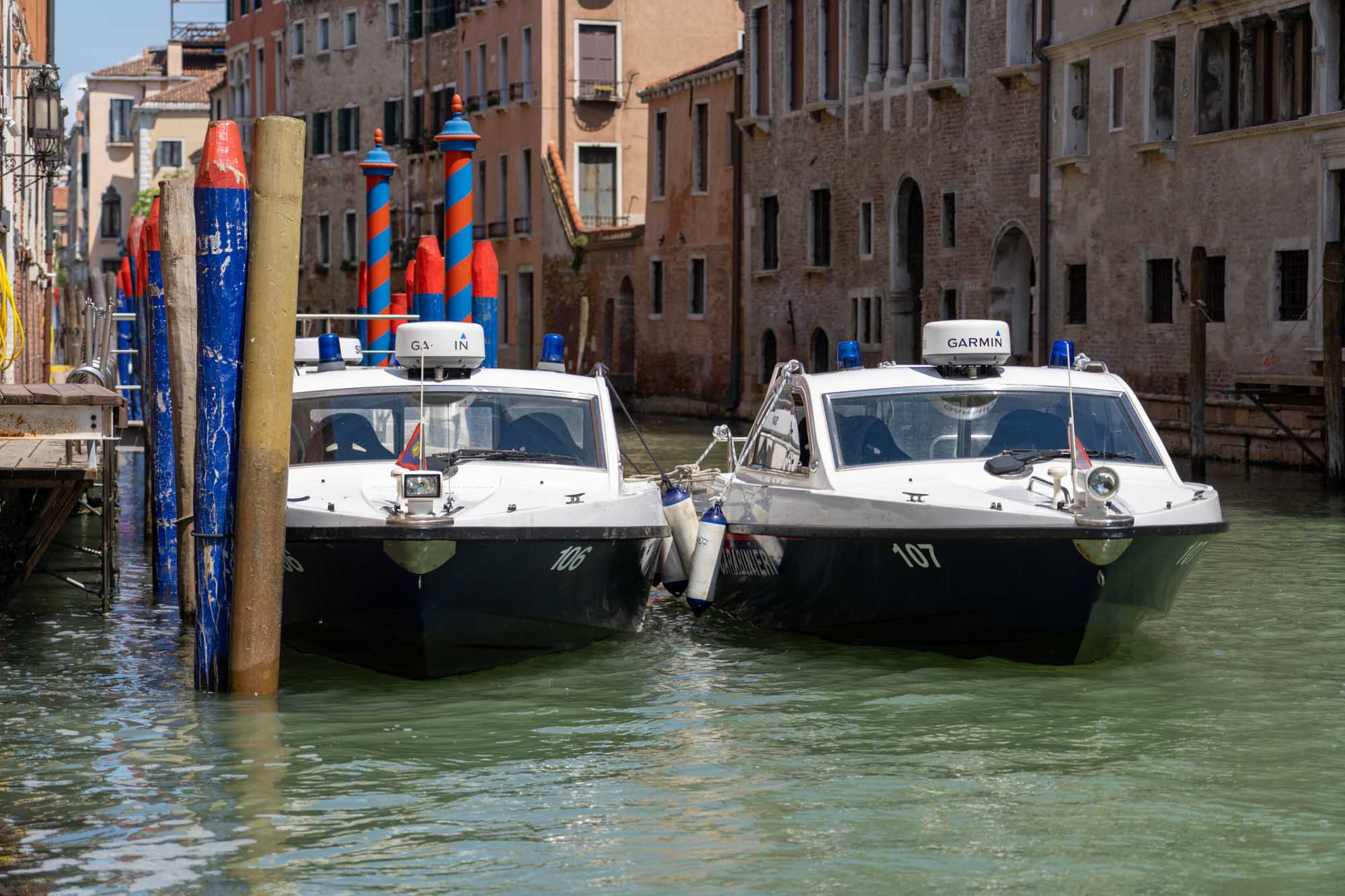 Two water taxis docked in a Venetian canal with colorful striped mooring poles and historic buildings in the background.