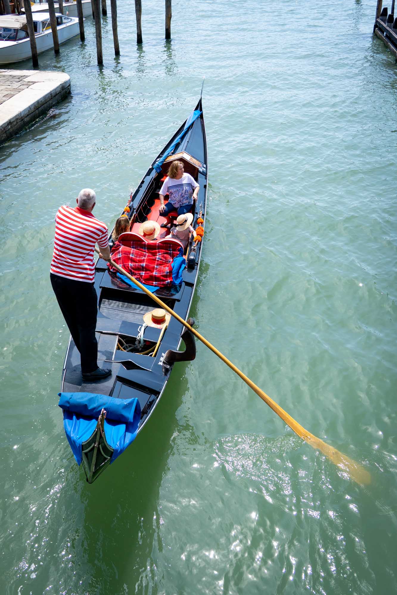 Gondolier in striped shirt rows tourists through Venice canal in gondola on sunny day.