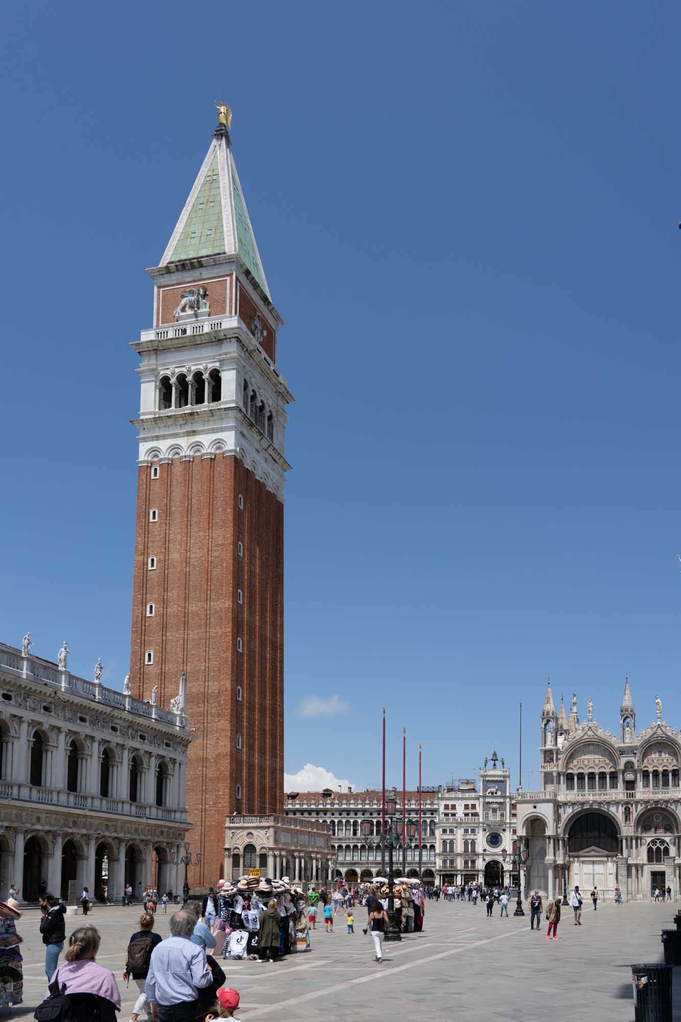 St. Mark's Campanile and Basilica in Venice, Italy with crowds in Piazza San Marco under a clear blue sky.