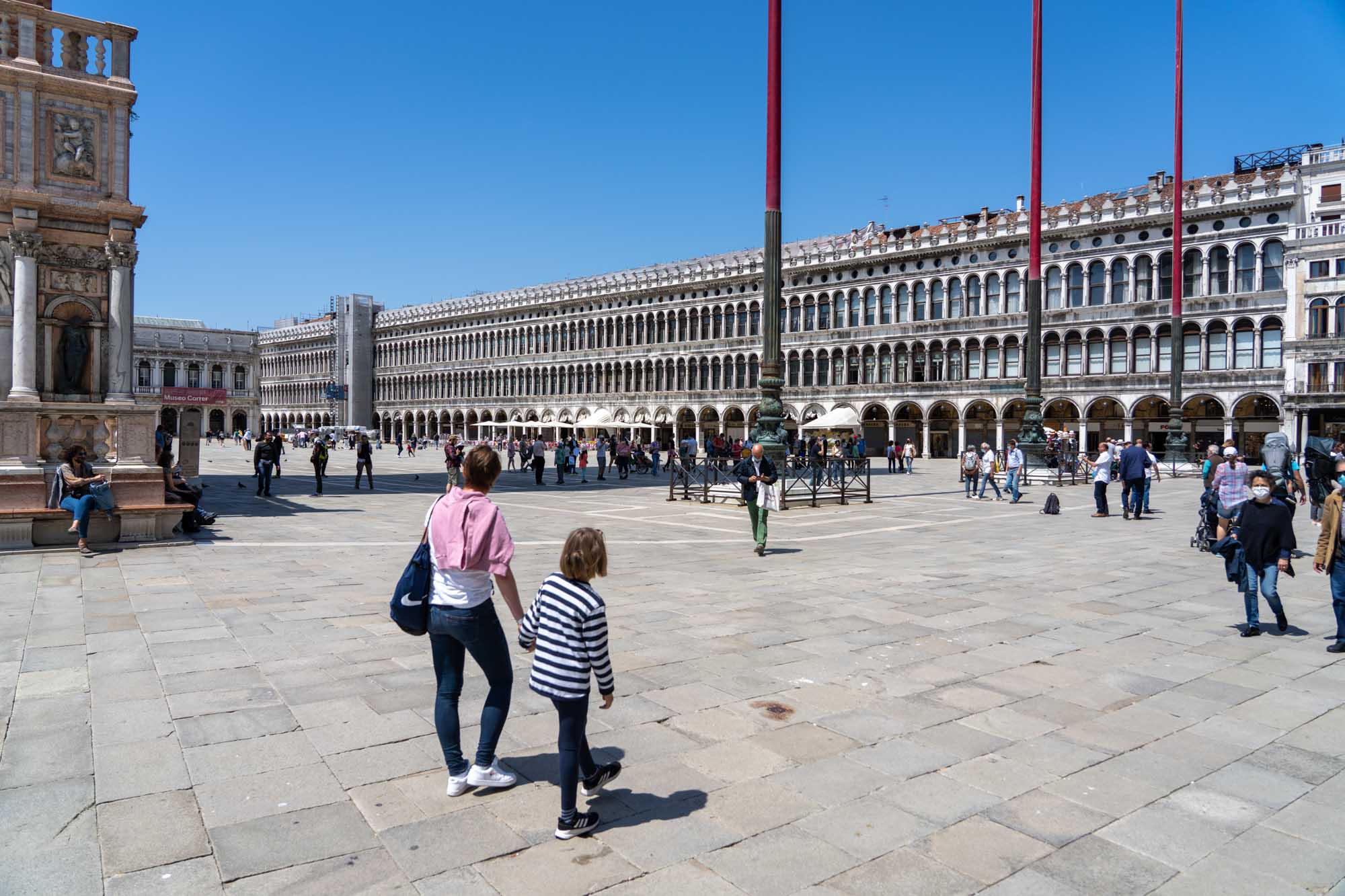 People walking in St. Mark's Square, Venice, with historic buildings and a clear blue sky in the background.