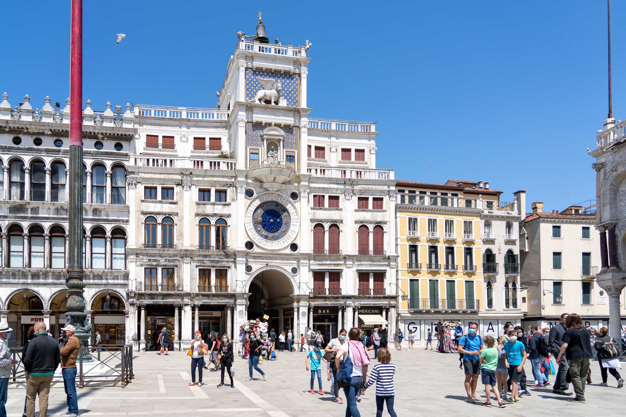 People explore St. Mark's Clock Tower plaza in Venice, Italy, under a clear blue sky.