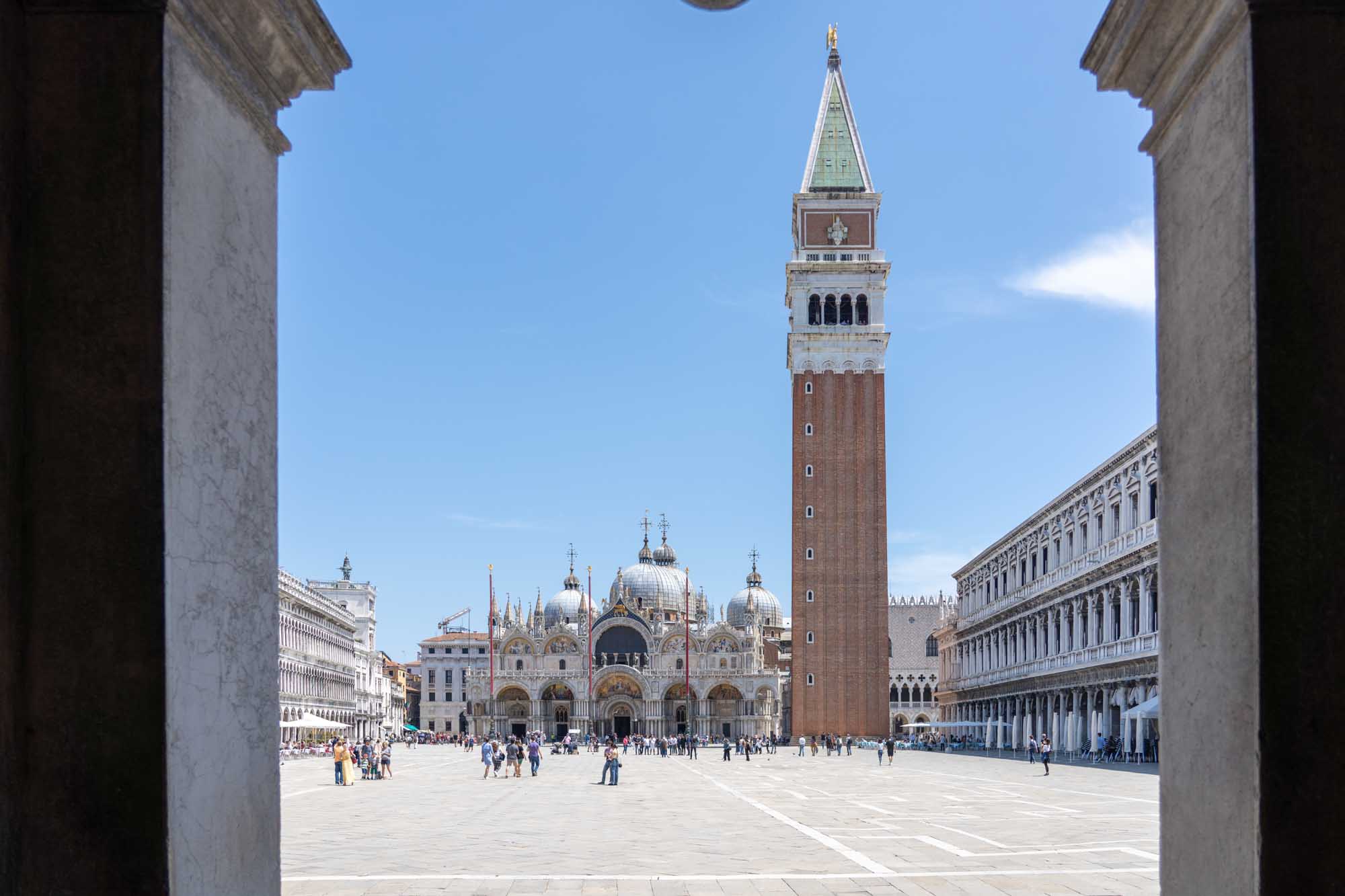 View of St. Mark's Basilica and Campanile tower in Venice, framed by archway, with tourists on sunny day.
