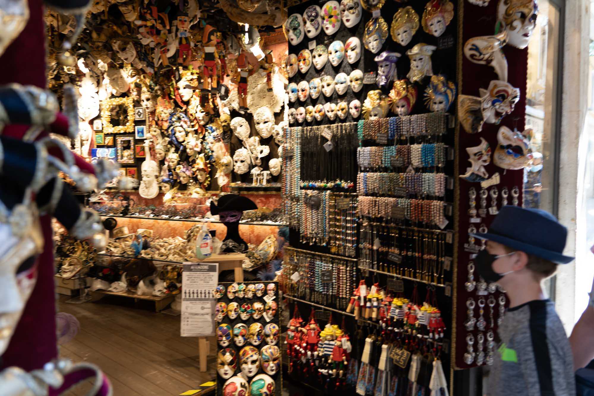 Venetian mask shop filled with colorful masks and jewelry, a person in a hat and mask walks by the vibrant display.