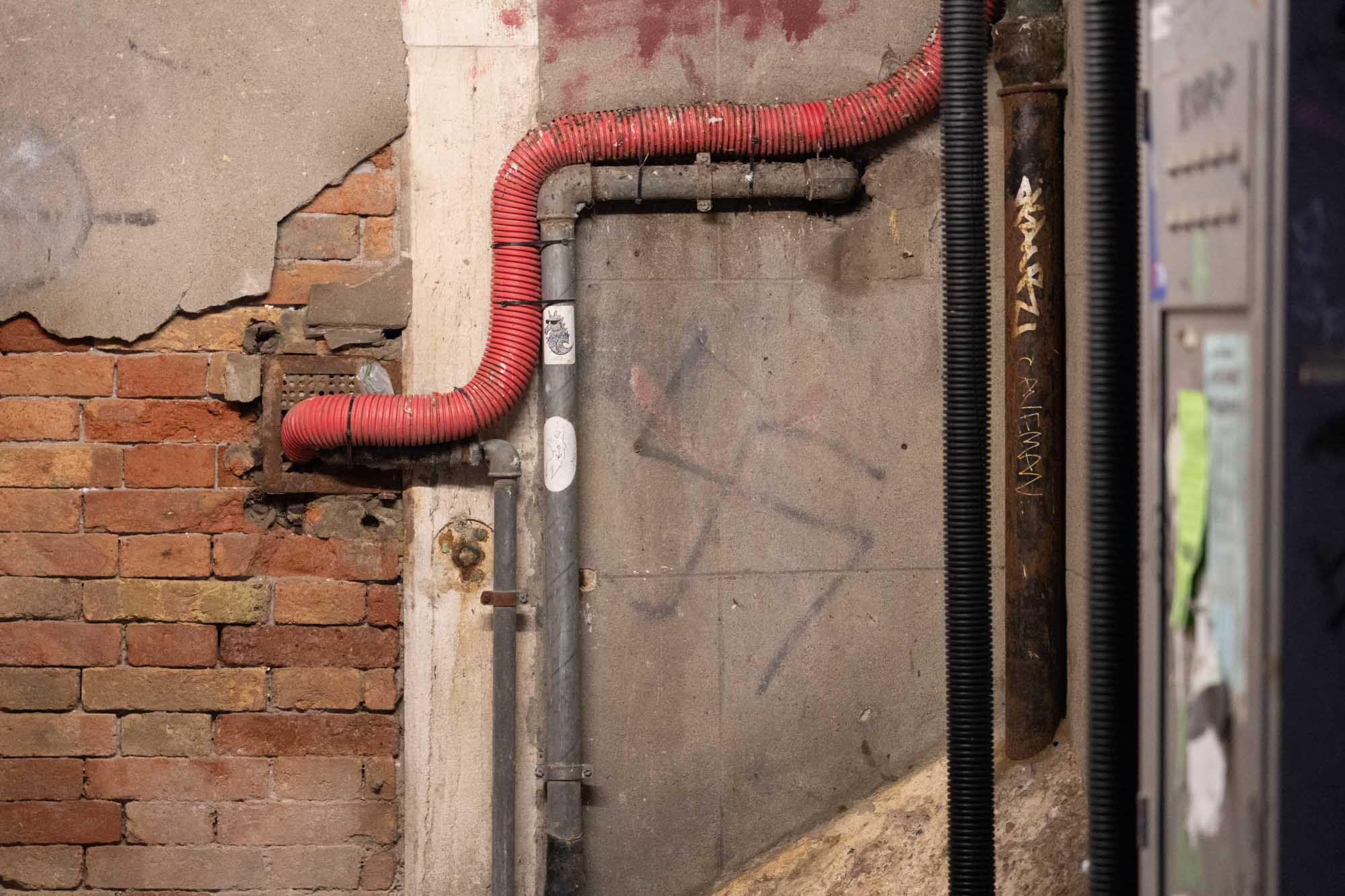 Old brick wall with red and black pipes, graffiti, and exposed concrete, showcasing an urban, industrial aesthetic.