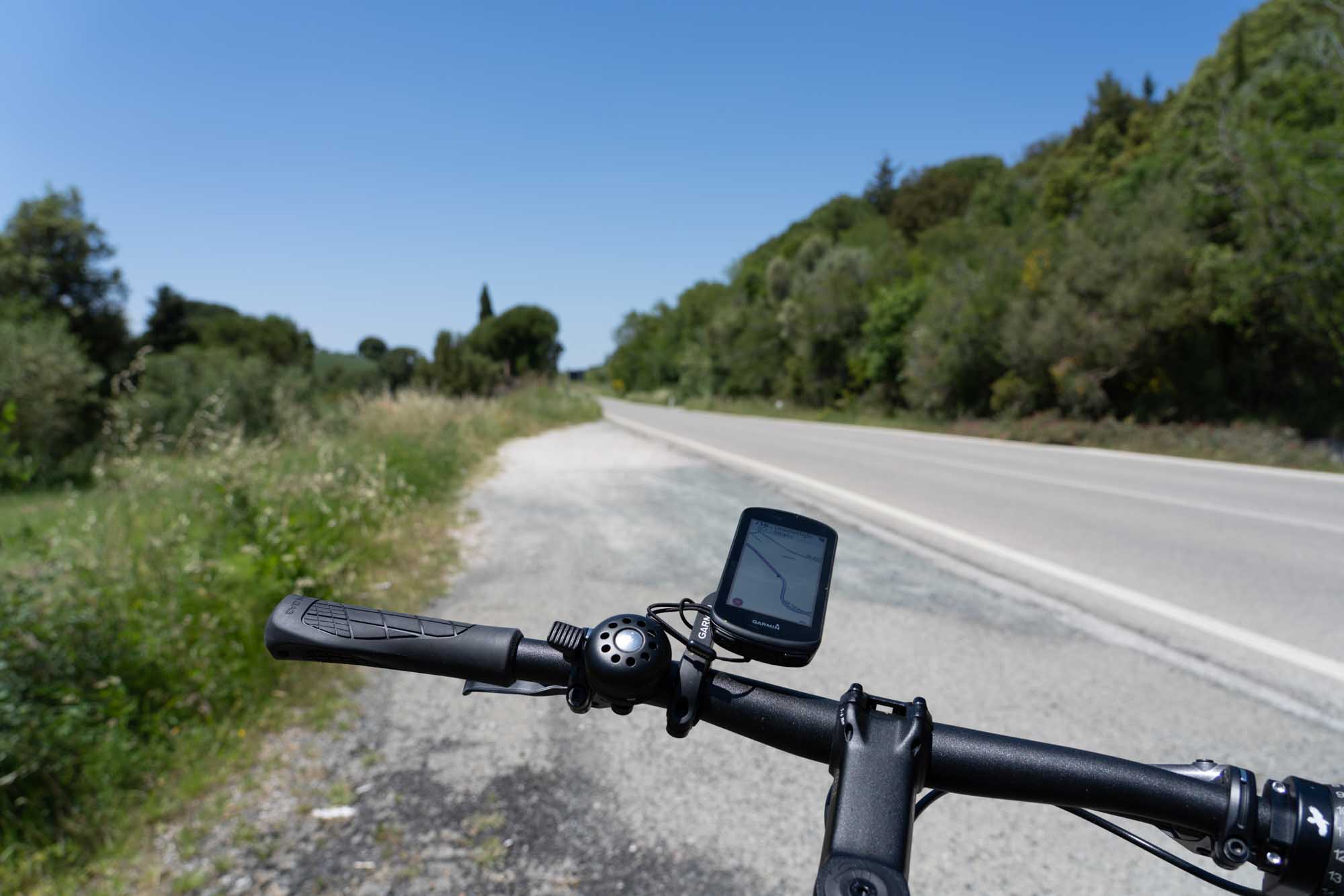 Bicycle handlebars with a GPS device on a scenic road bordered by greenery and a clear blue sky.
