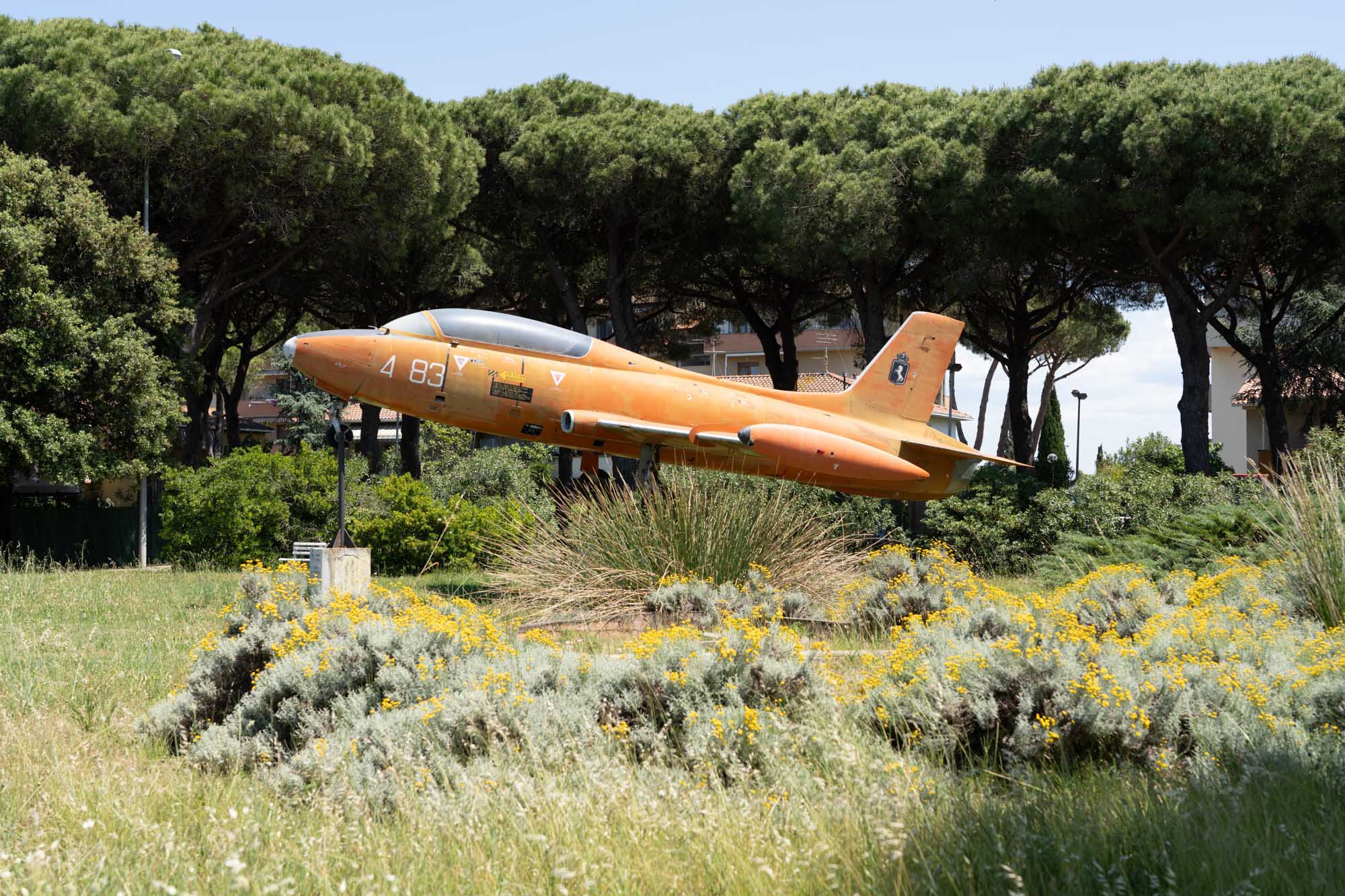 Vintage orange aircraft display in park, surrounded by lush greenery and wildflowers under a clear blue sky.