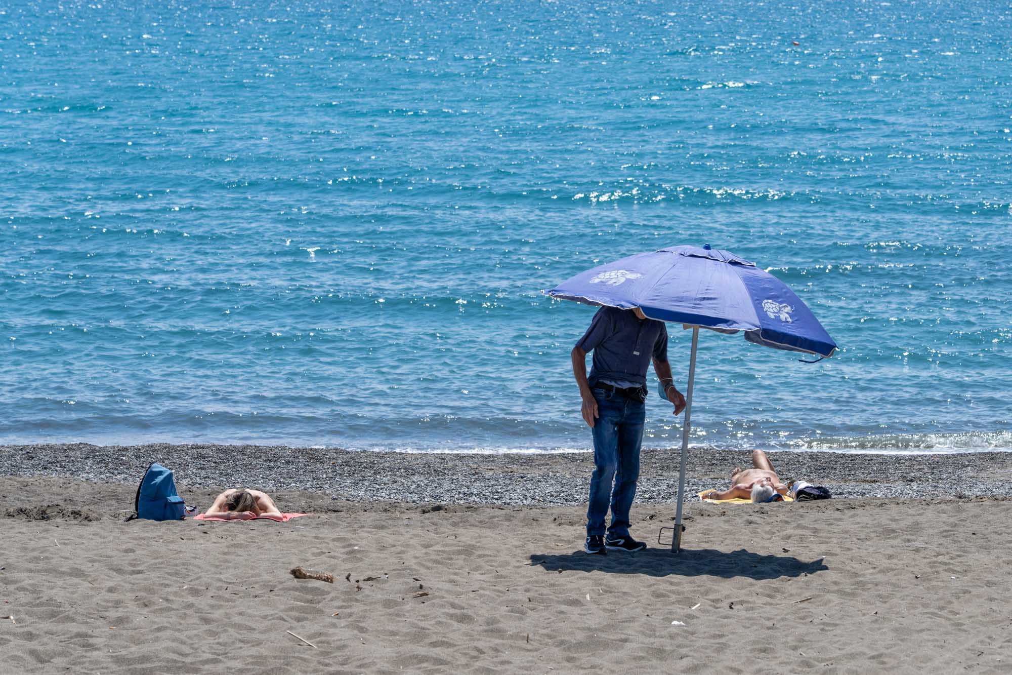 Man setting up blue umbrella on sandy beach with people sunbathing by the ocean.