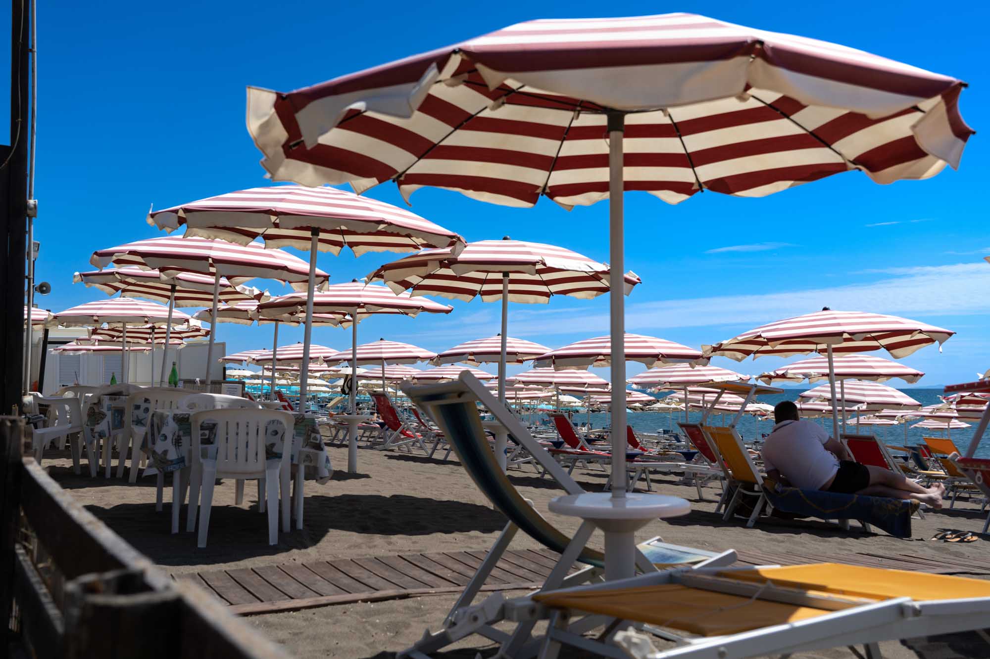 Beach scene with red and white umbrellas, lounge chairs, and a person relaxing under a clear blue sky near the ocean.