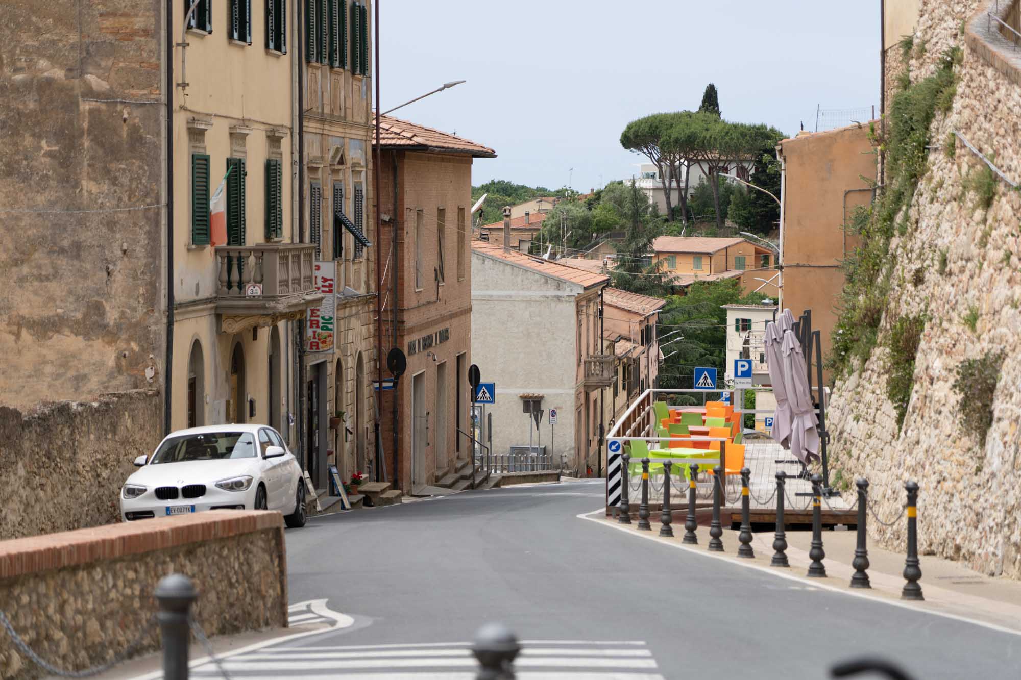Narrow Italian street with parked white car, colorful chairs, and historic buildings in countryside town.
