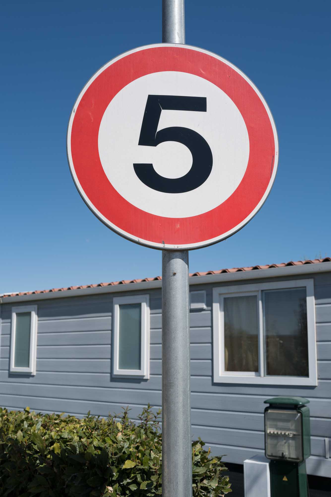 Circular speed limit sign with number 5 in front of a house and clear blue sky in the background.