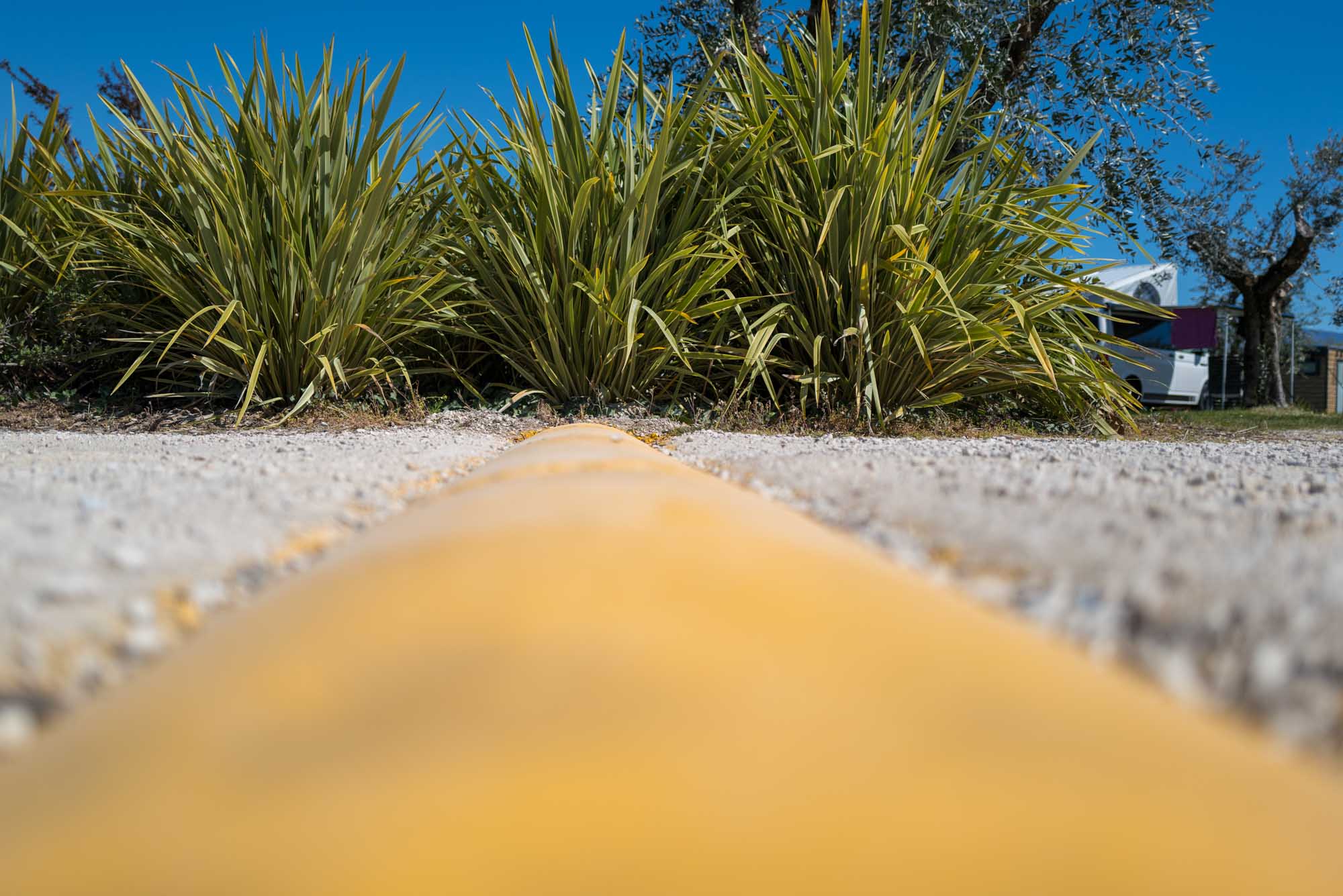 Close-up of a yellow speed bump on gravel, with green bushes and a white camper van in the background, against a clear blue sky.