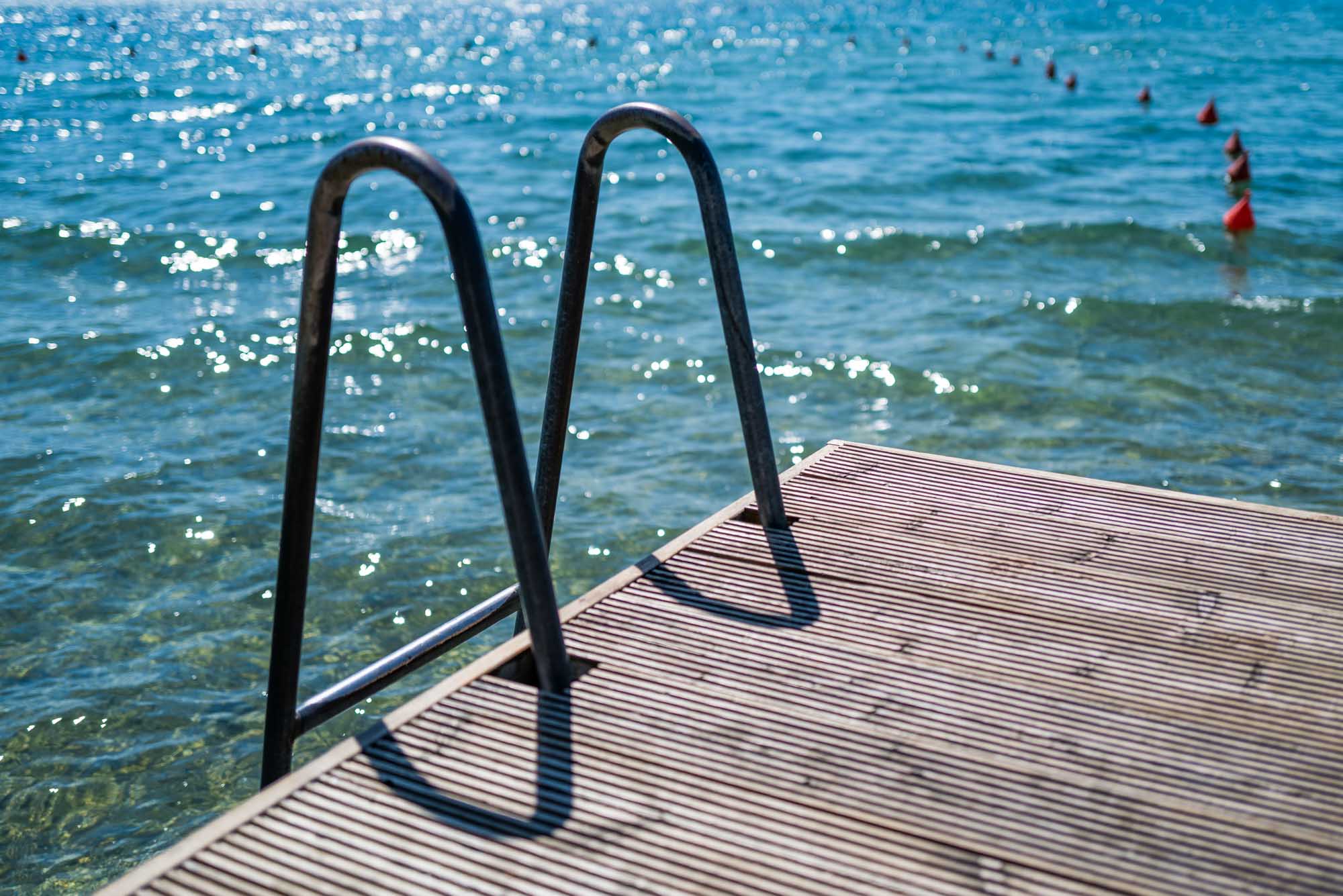 Ladder on a dock leading into clear blue lake water under a sunny sky with glistening waves and buoys in view.