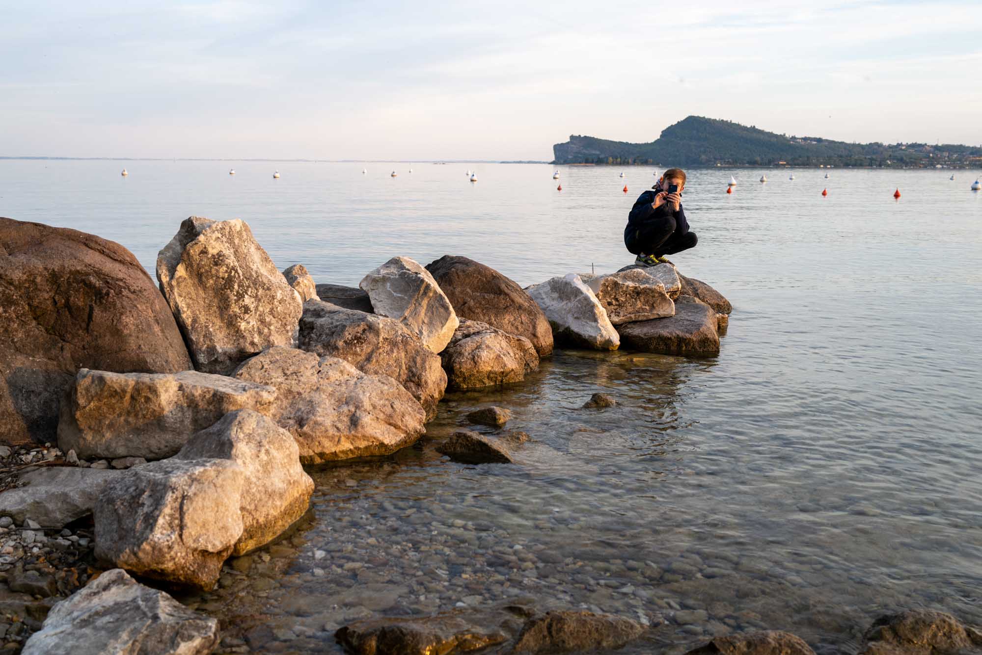Person crouching on rocks by a serene lake with distant hills and buoys in the background.