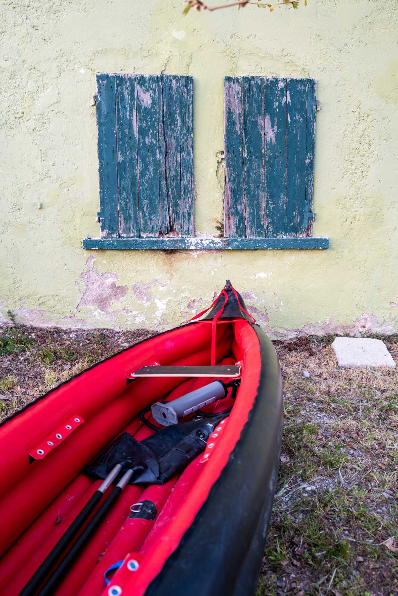 Red kayak with paddles and gear against an old wall with weathered green shutters, showcasing rustic outdoor adventure.