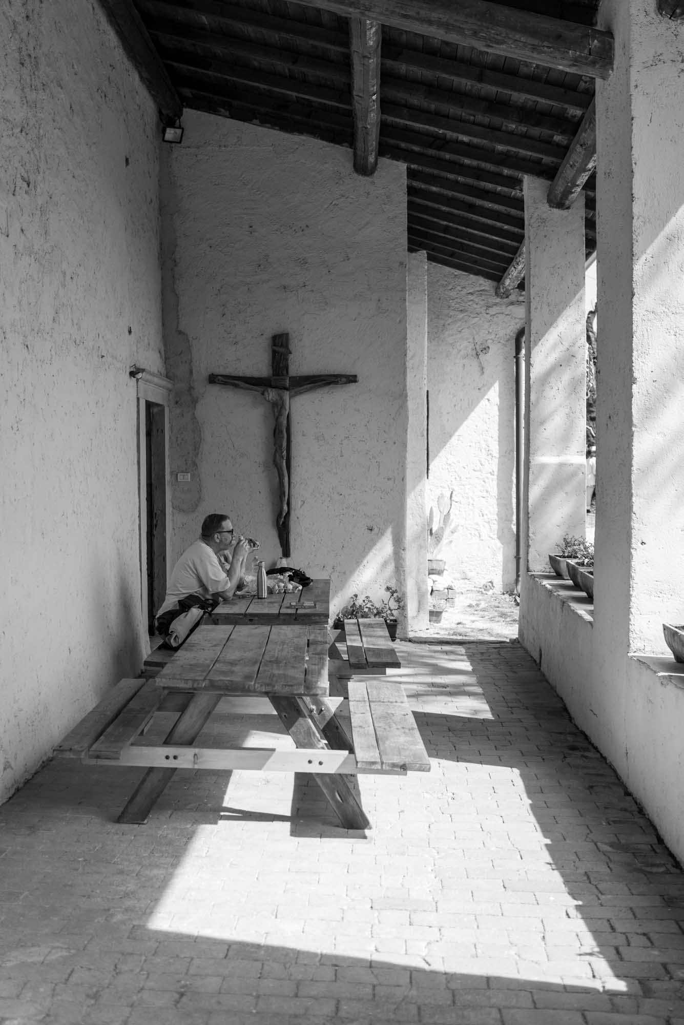 Man sitting at a picnic table under a cross, in a shaded rustic corridor with sunlight streaming, captured in black and white.