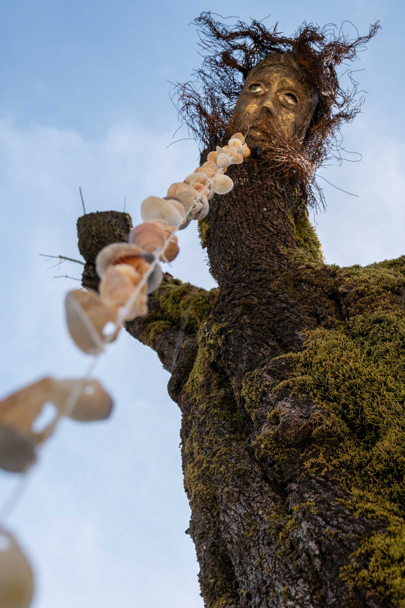 Tree sculpture with mossy bark, featuring a face and shell garland, set against a blue sky.