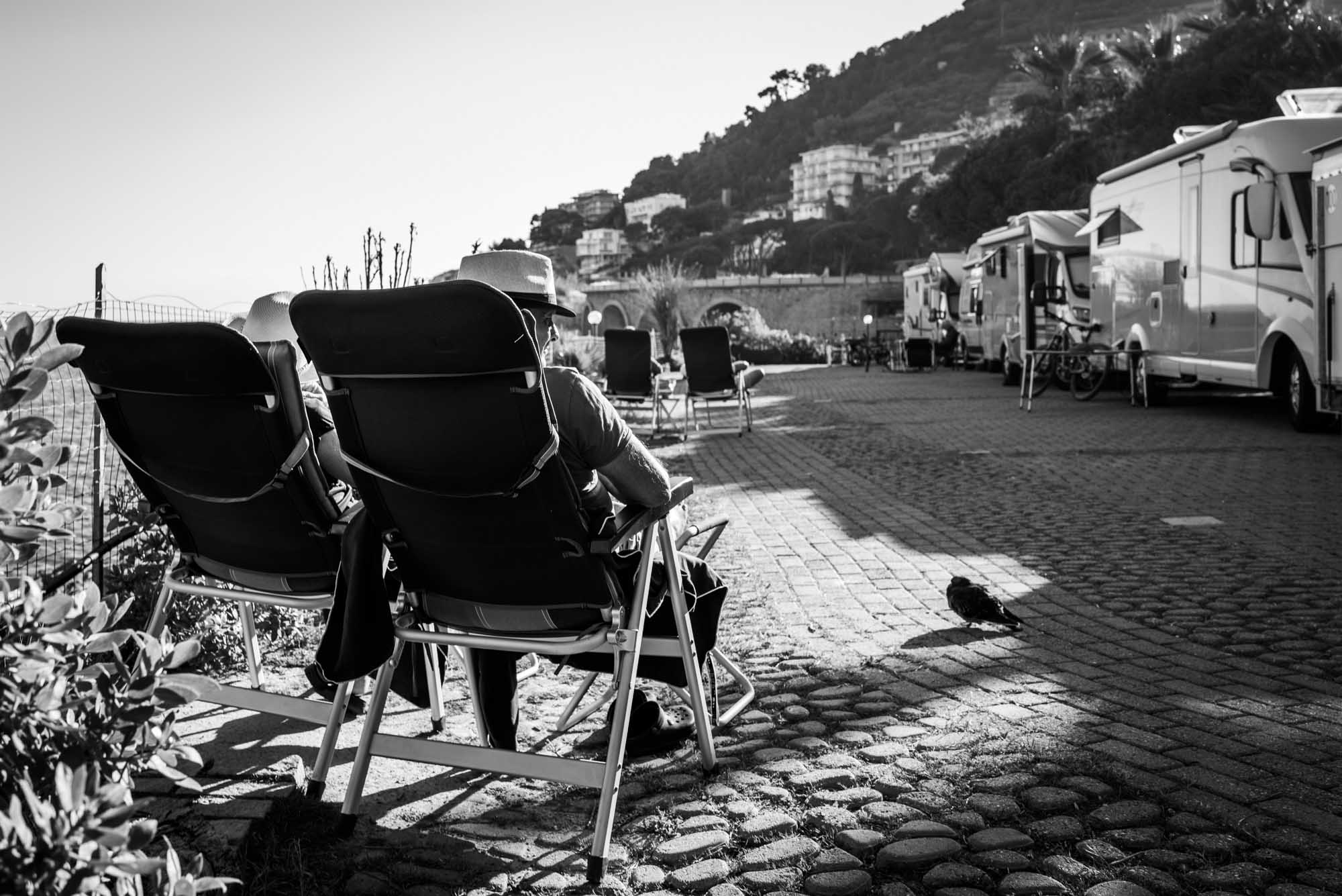 Two people in hats relax on lounge chairs by a coastal RV park, with scenic hills and buildings in the background.