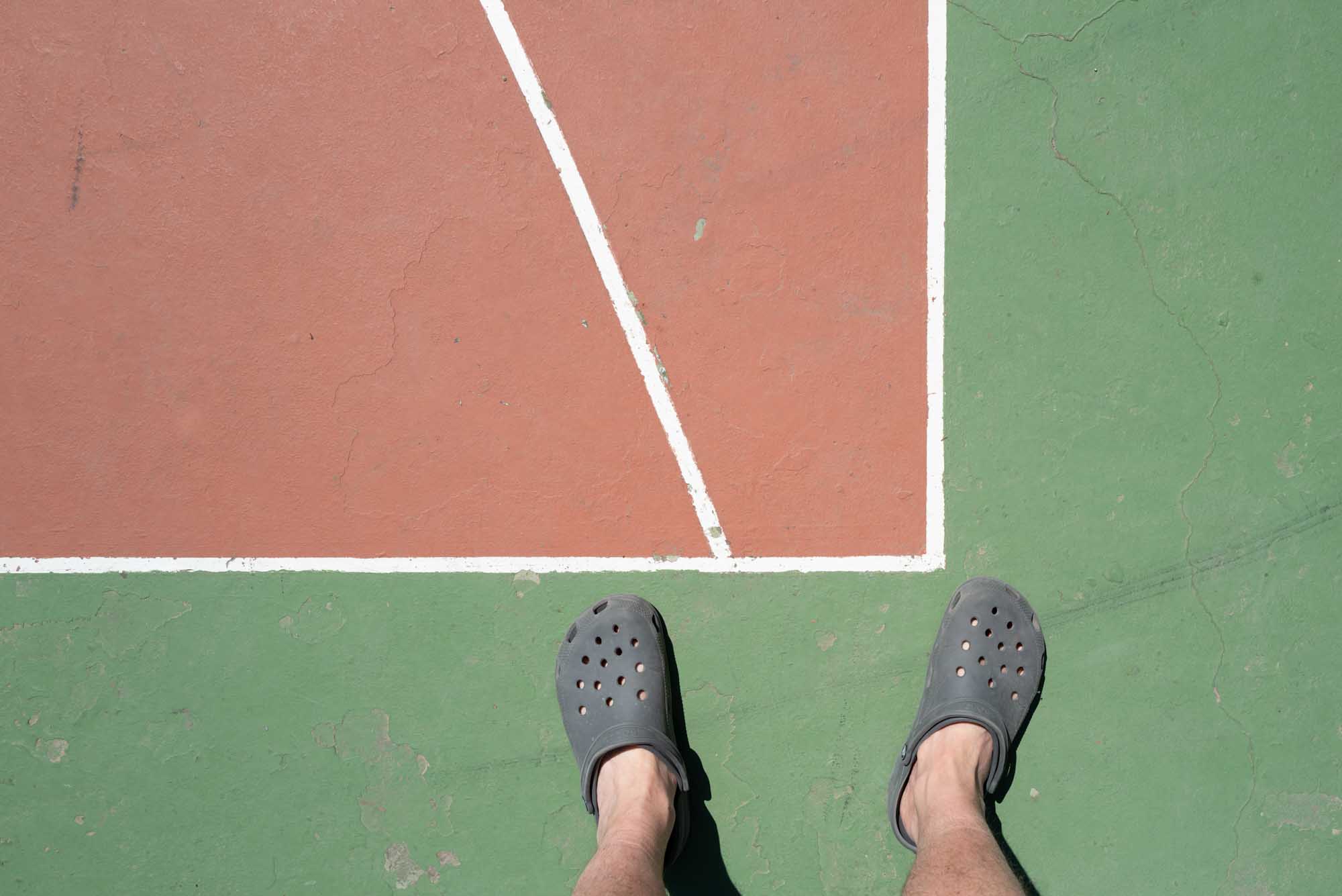 Pair of feet in casual clogs standing on a green and red sports court with white lines.