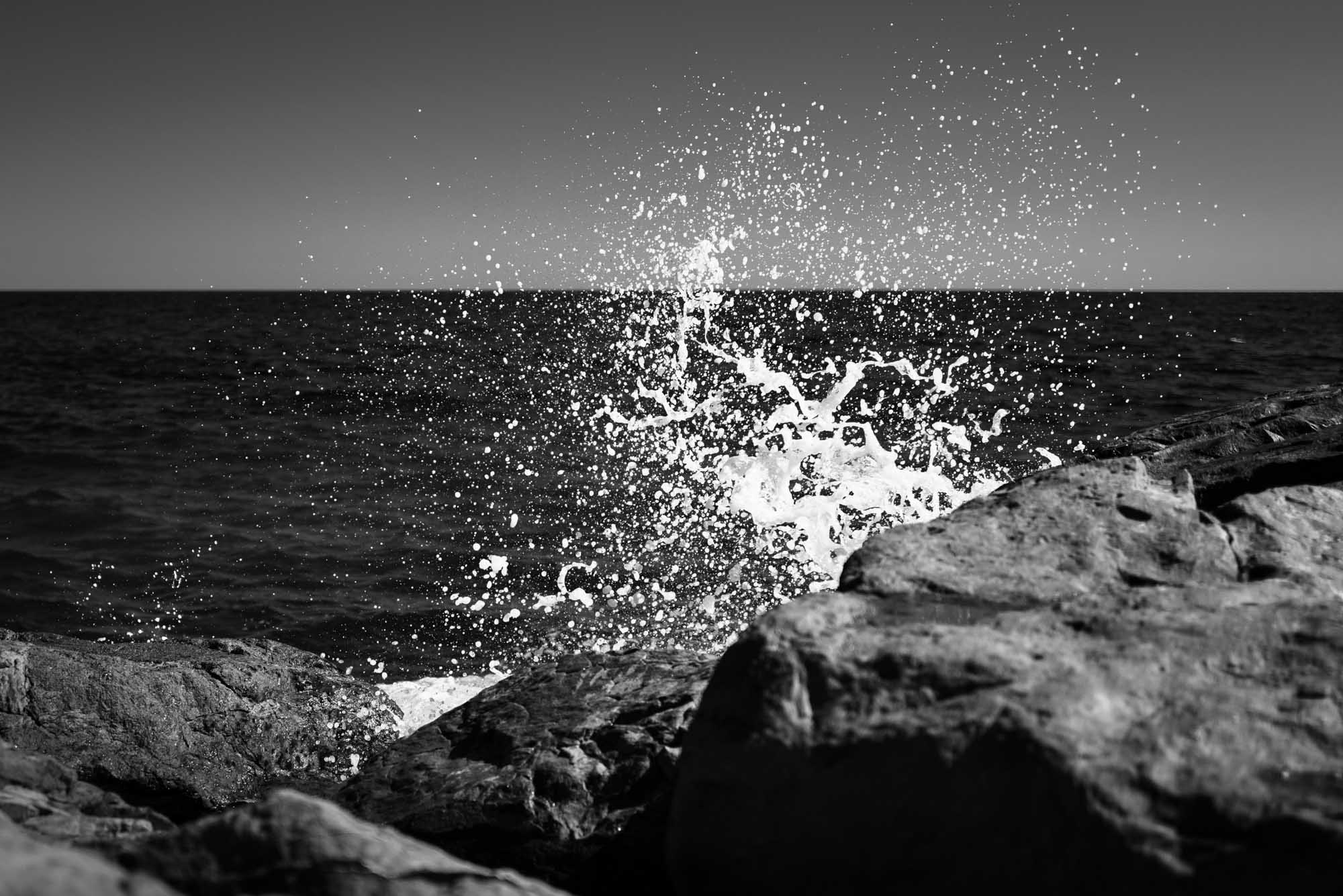 Waves crashing against rocks, creating a splash under a clear sky in a black and white photograph.