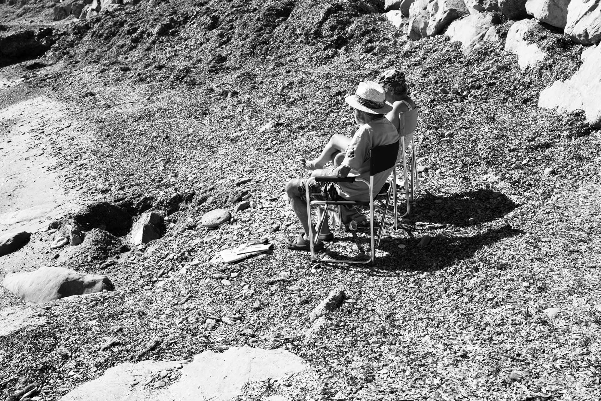 Two people seated on rocky ground, wearing hats, in a sunlit outdoor setting.