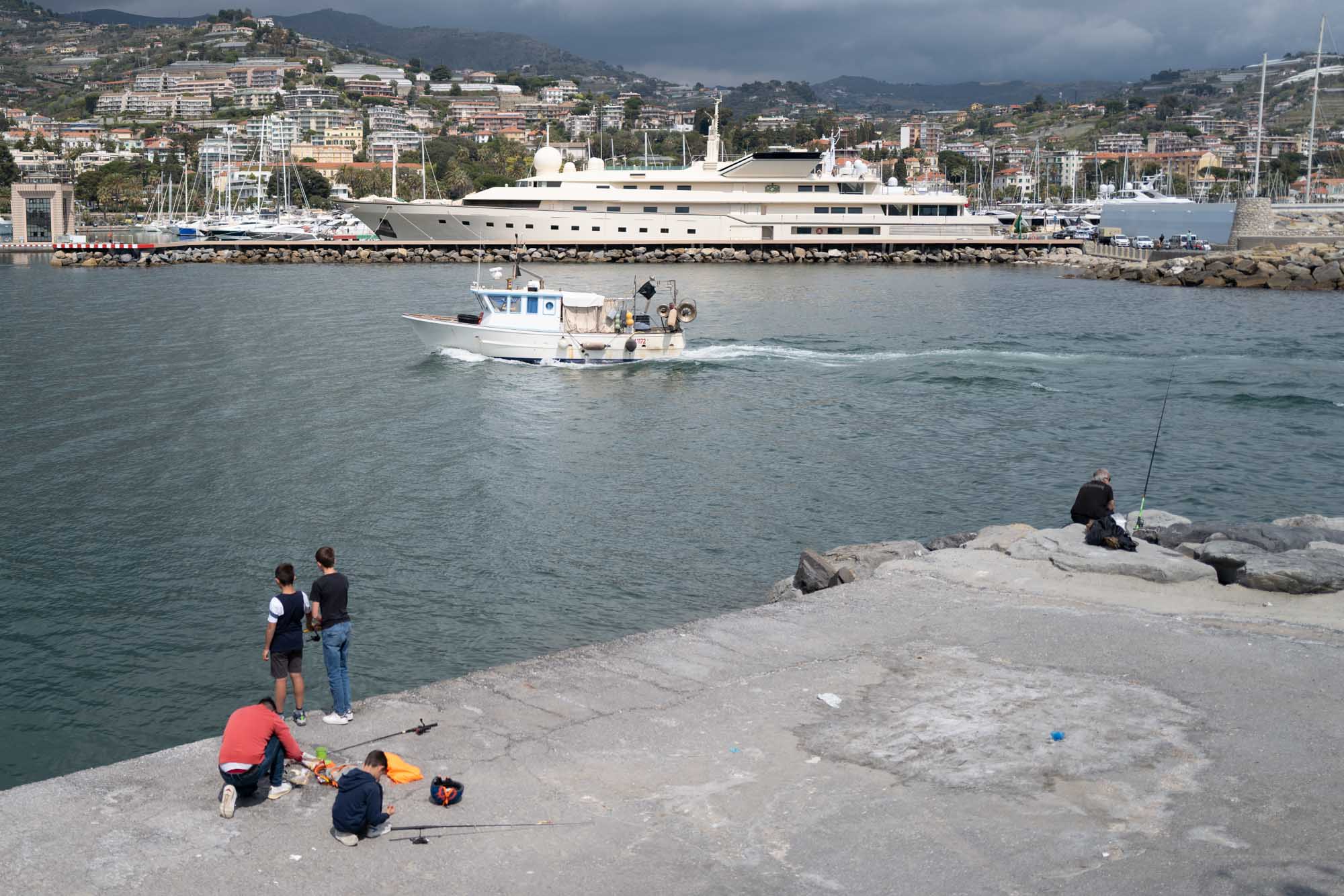 People fishing on a pier watch a small boat pass by a large yacht in a harbor, with hills and buildings in the background.