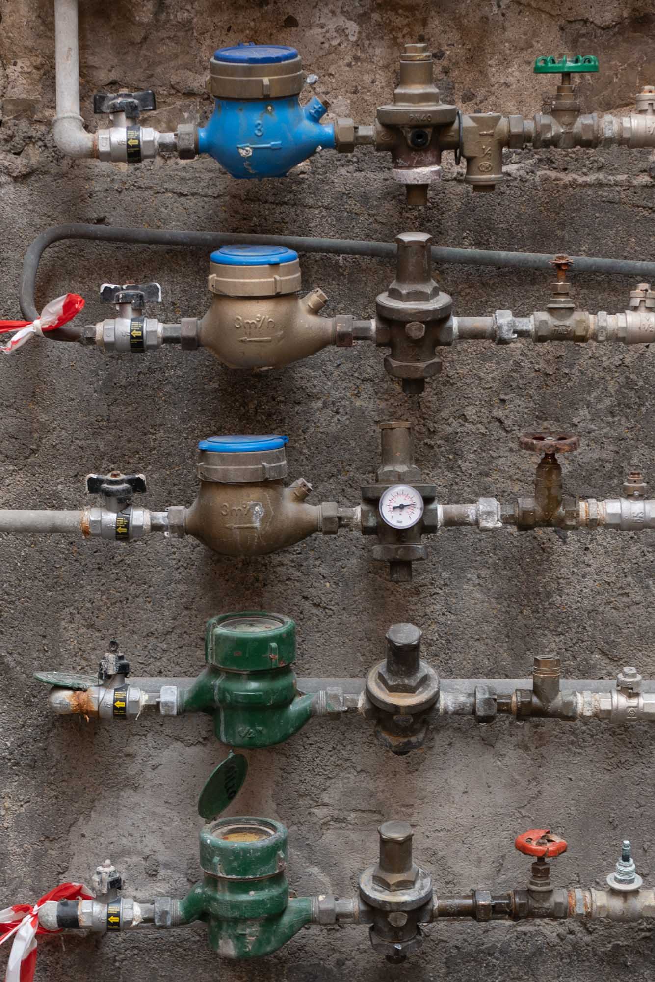 Rows of colorful water meters and valves on a textured wall, featuring blue and green gauges with pipes and connections.