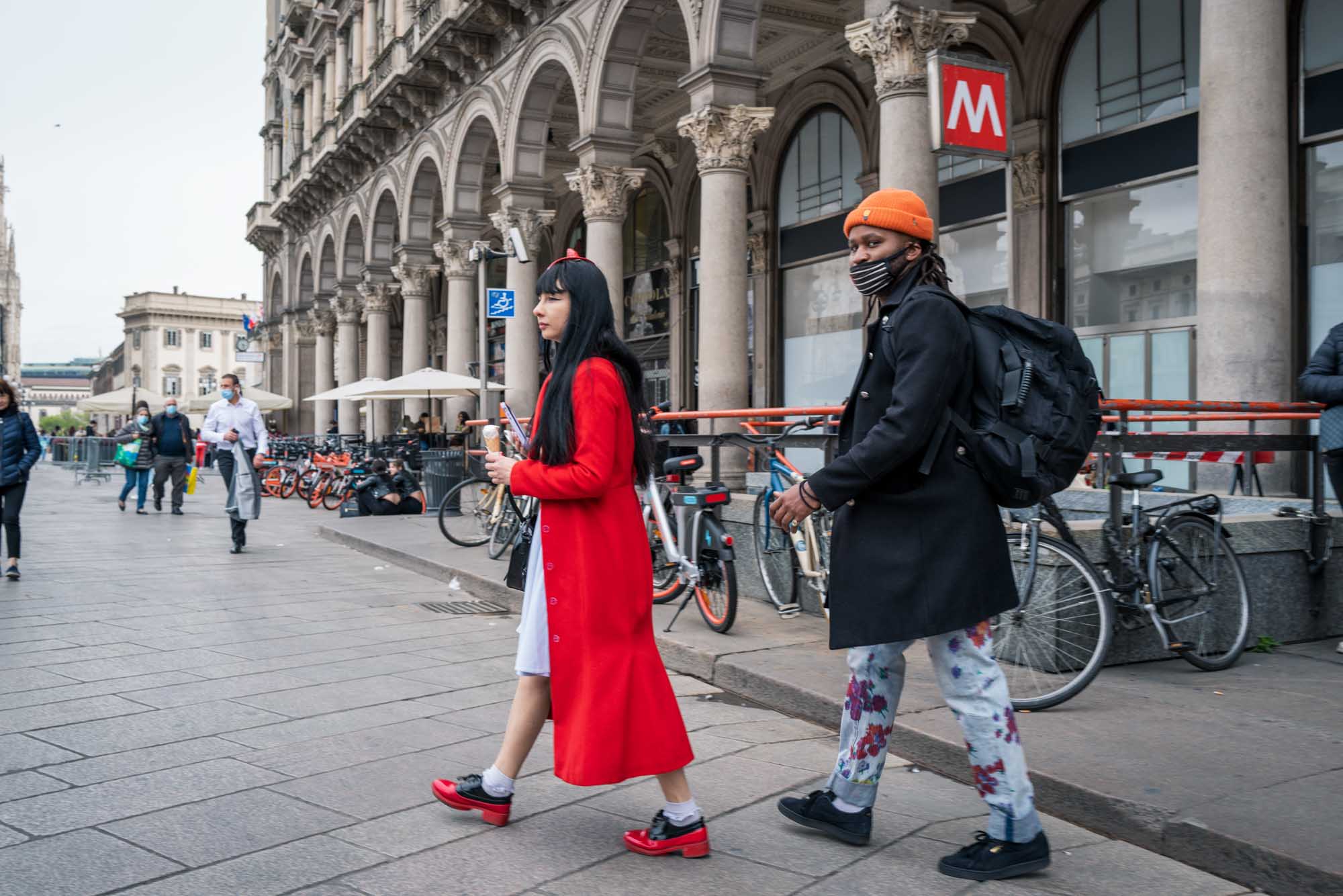 Two people walking past bikes and a metro station in front of an ornate building, one in a red coat, the other in an orange beanie.