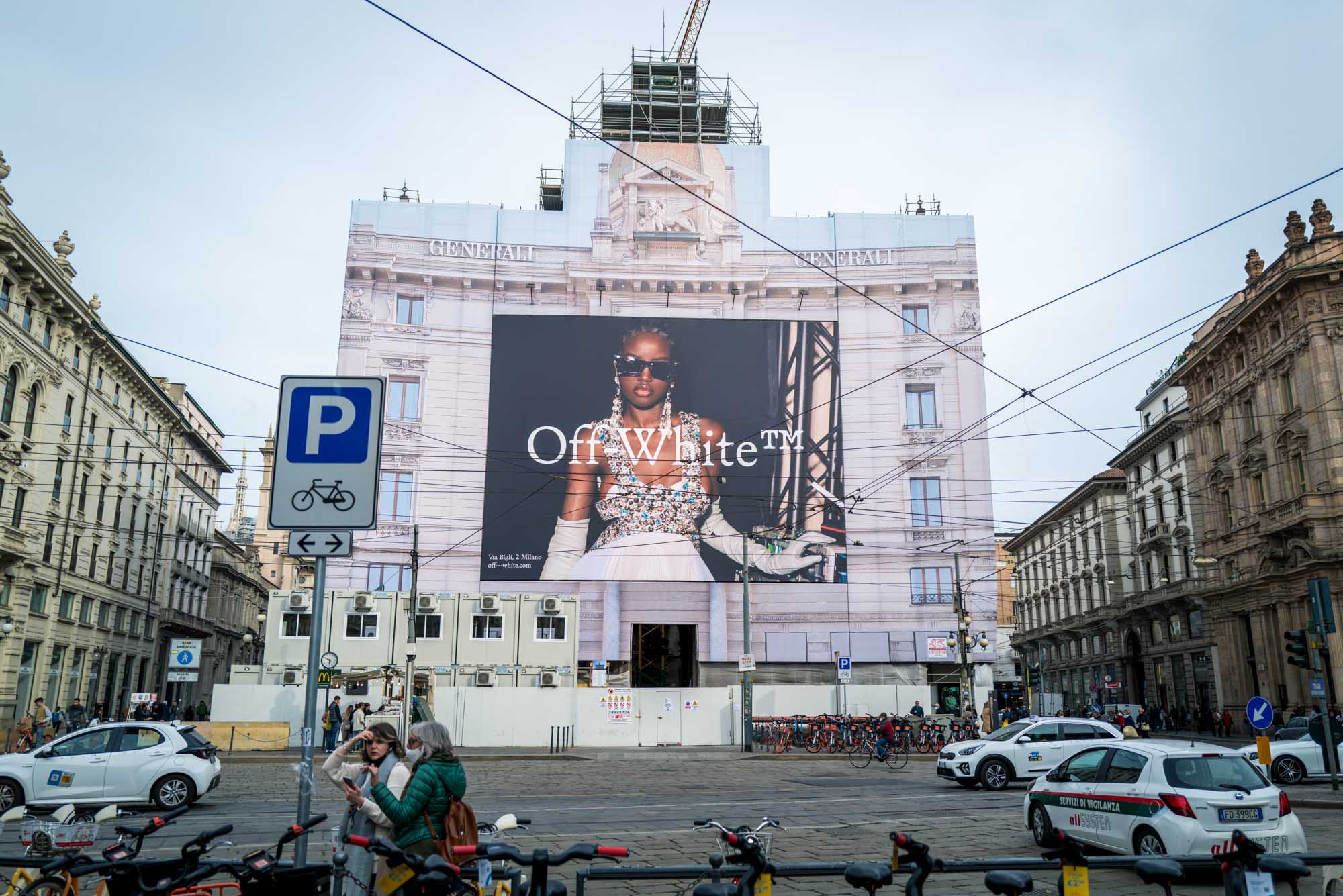 Large Off-White ad covering building facade in bustling city intersection with bikes, taxis, and pedestrians.